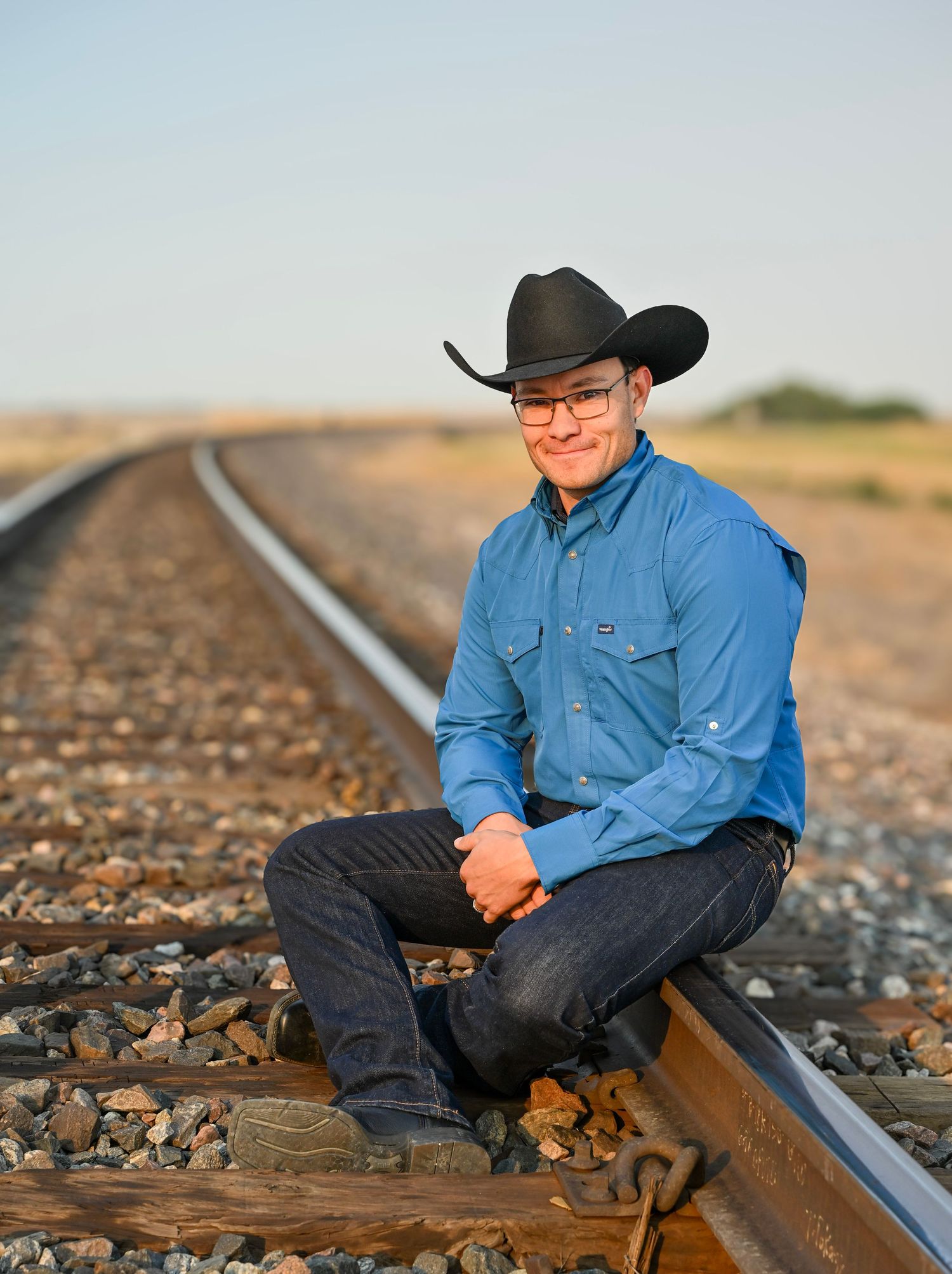 Young man in black cowboy hat and blue shirt sitting on railroad tracks in open plains.