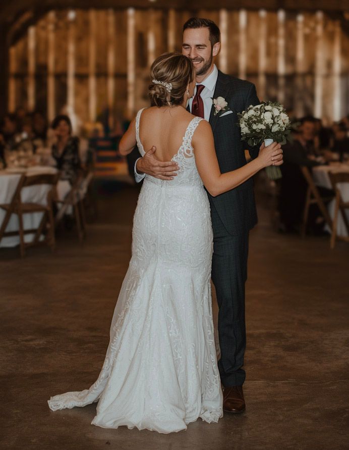 Bride and groom sharing first dance in rustic barn venue, bride wearing backless lace wedding gown.
