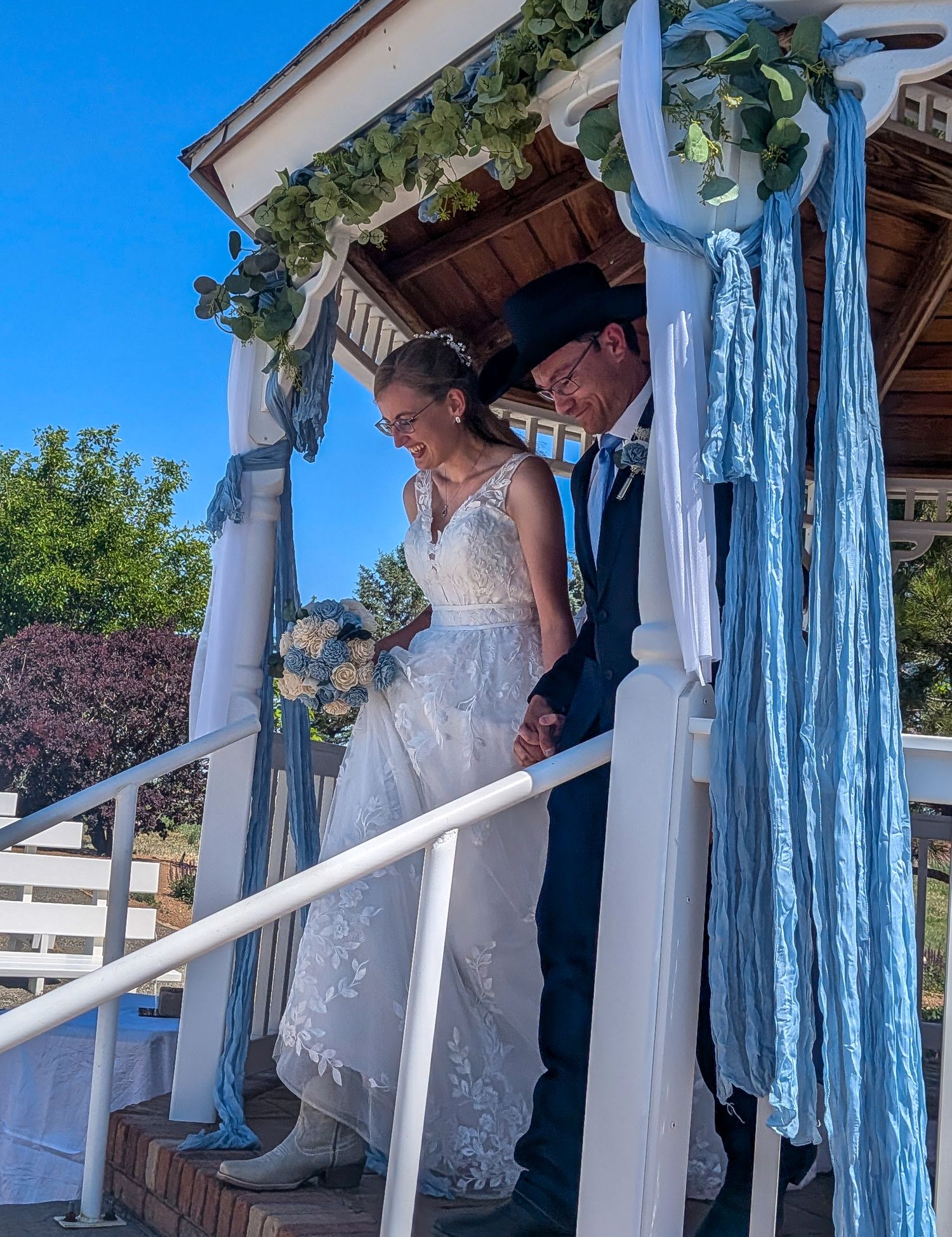 Bride and groom descending gazebo steps decorated with blue ribbons and greenery.