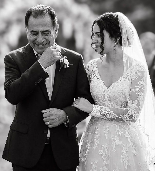 Father kissing bride's hand while she smiles on her wedding day, black and white photo.