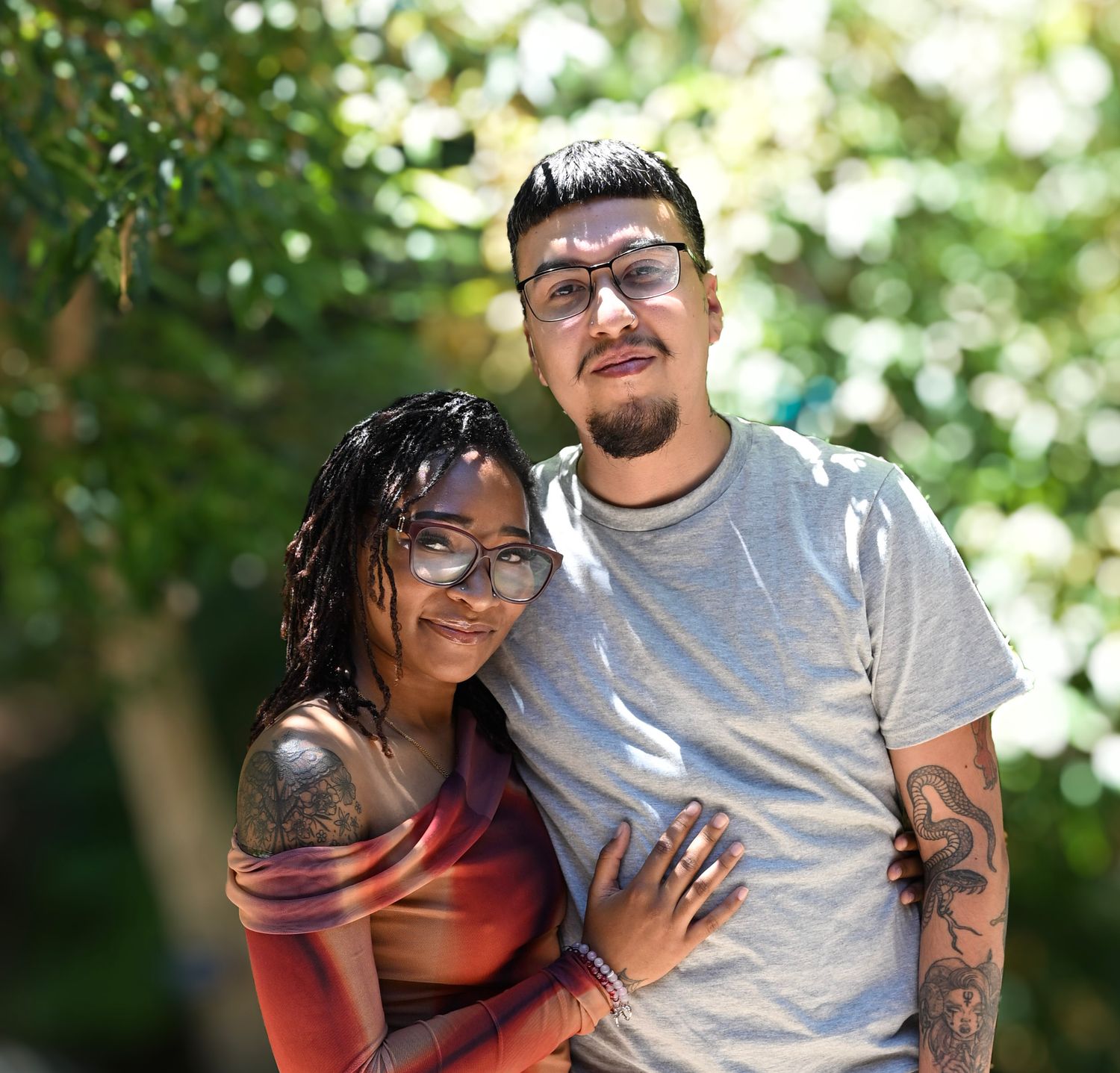 A smiling couple poses outdoors, woman with dreadlocks and man with glasses, both with tattoos, surrounded by green trees.