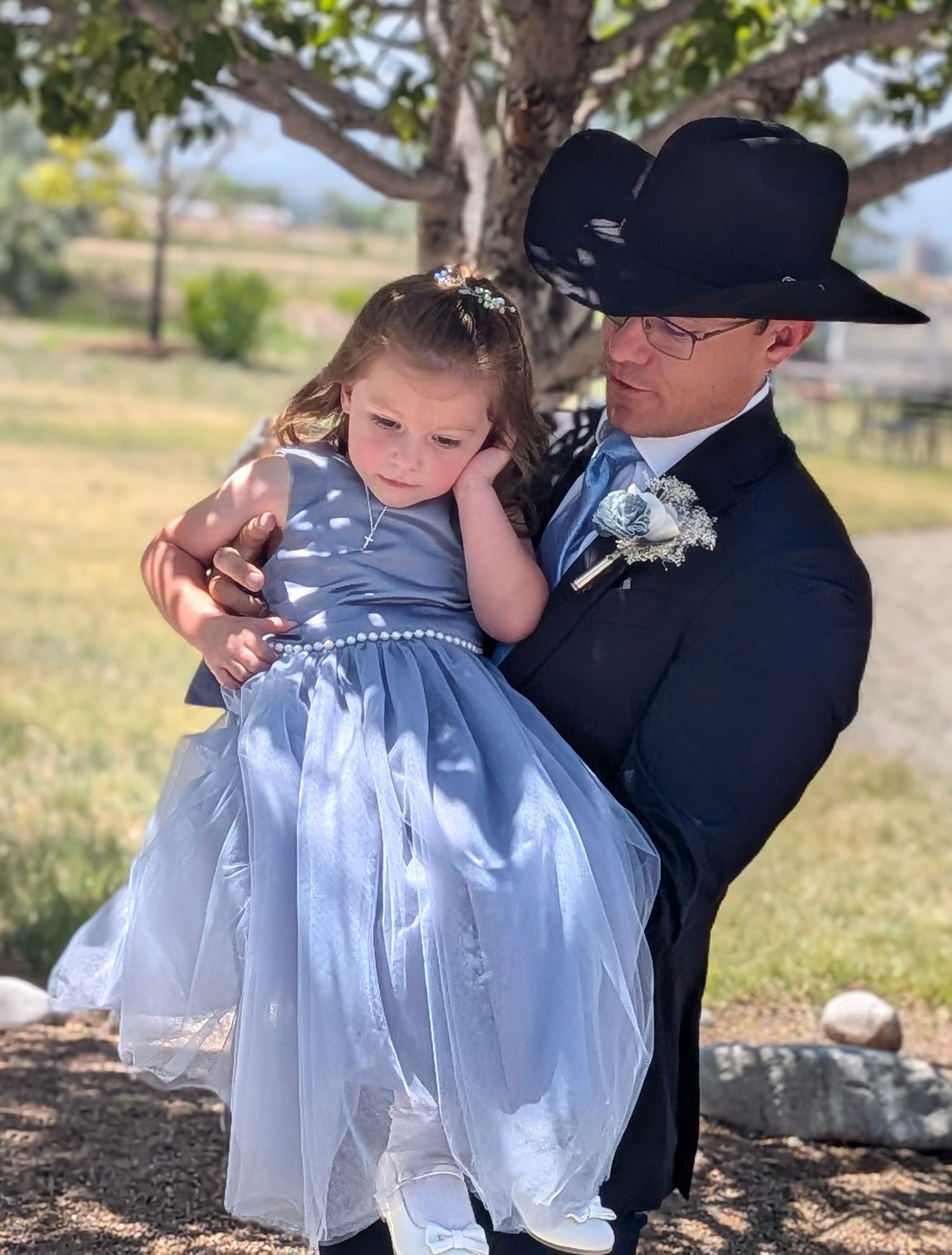 Groom in black suit and cowboy hat holding young flower girl in blue dress at outdoor wedding.