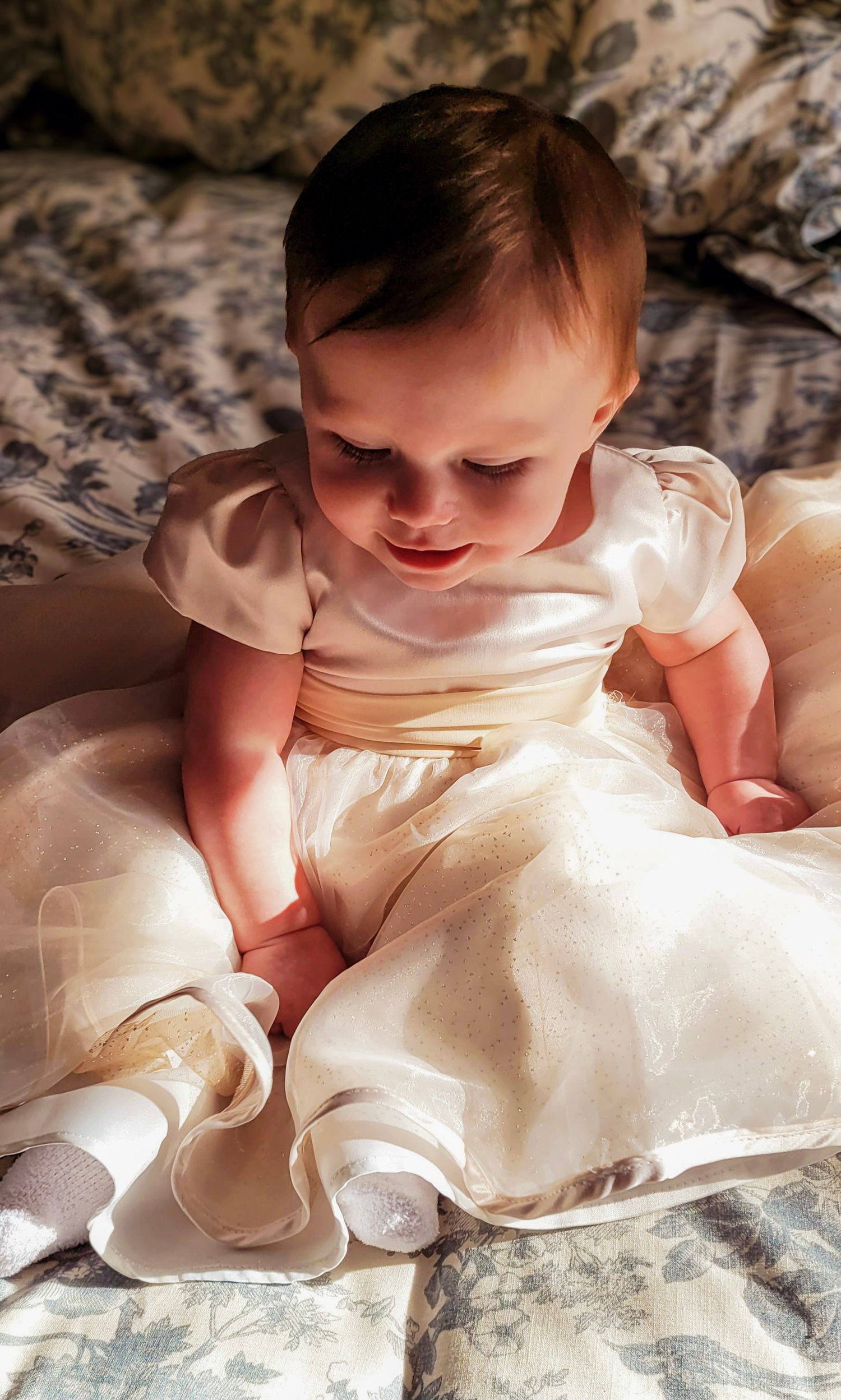 Adorable baby girl in white satin christening gown sitting on floral patterned bedding.