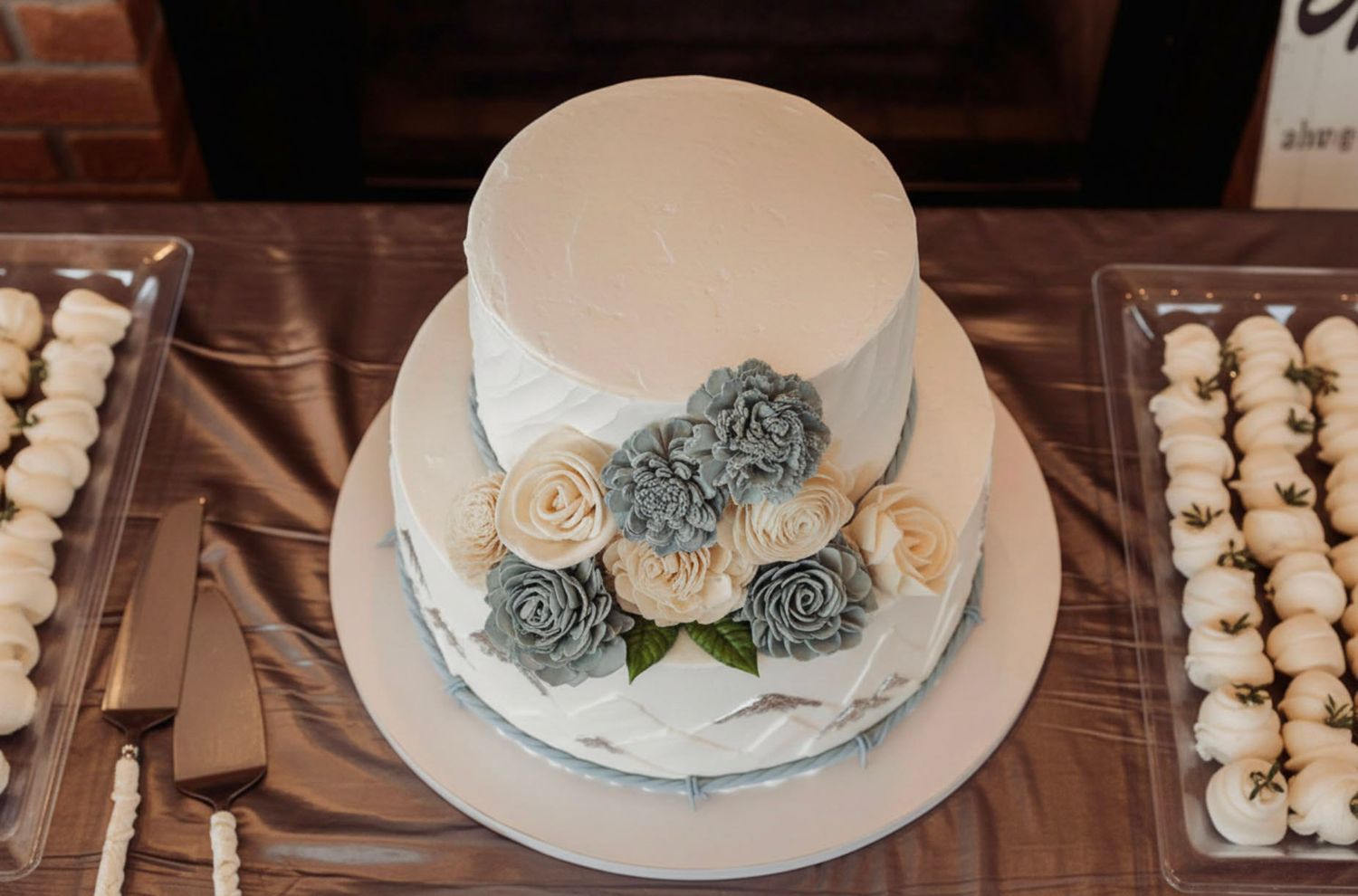 Two-tier white wedding cake decorated with blue and cream rosette flowers on a dessert table.