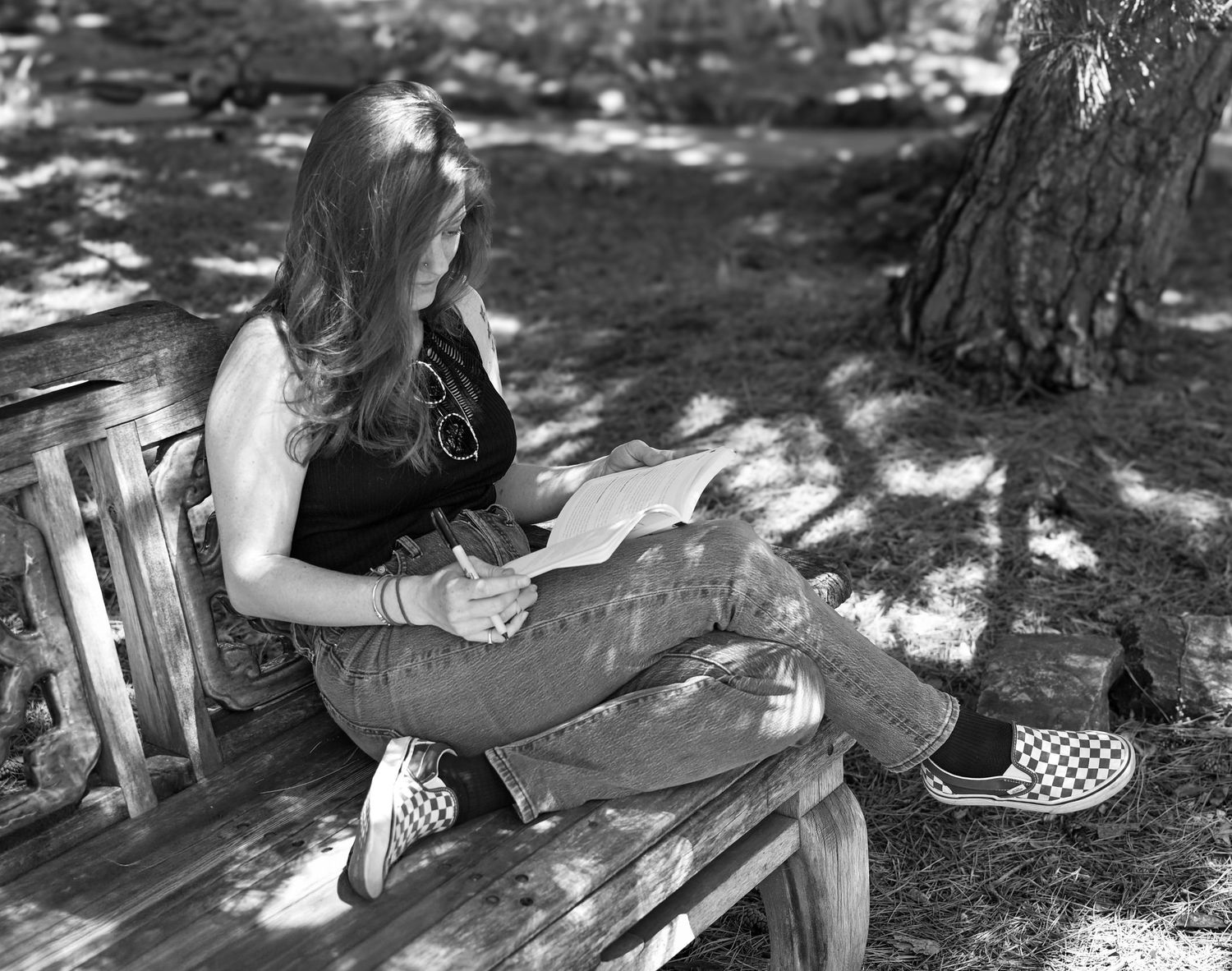 Woman reading a book on a wooden bench under a tree in black and white photo outdoors.