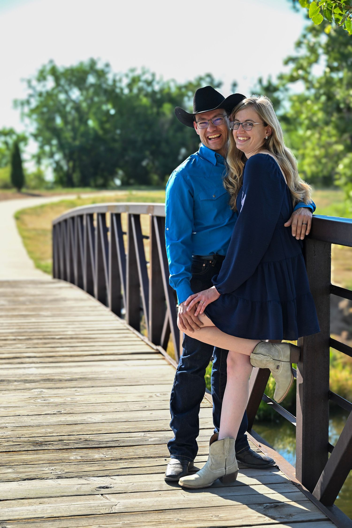 Happy couple in matching blue outfits and cowboy hats pose on a wooden bridge in a park.