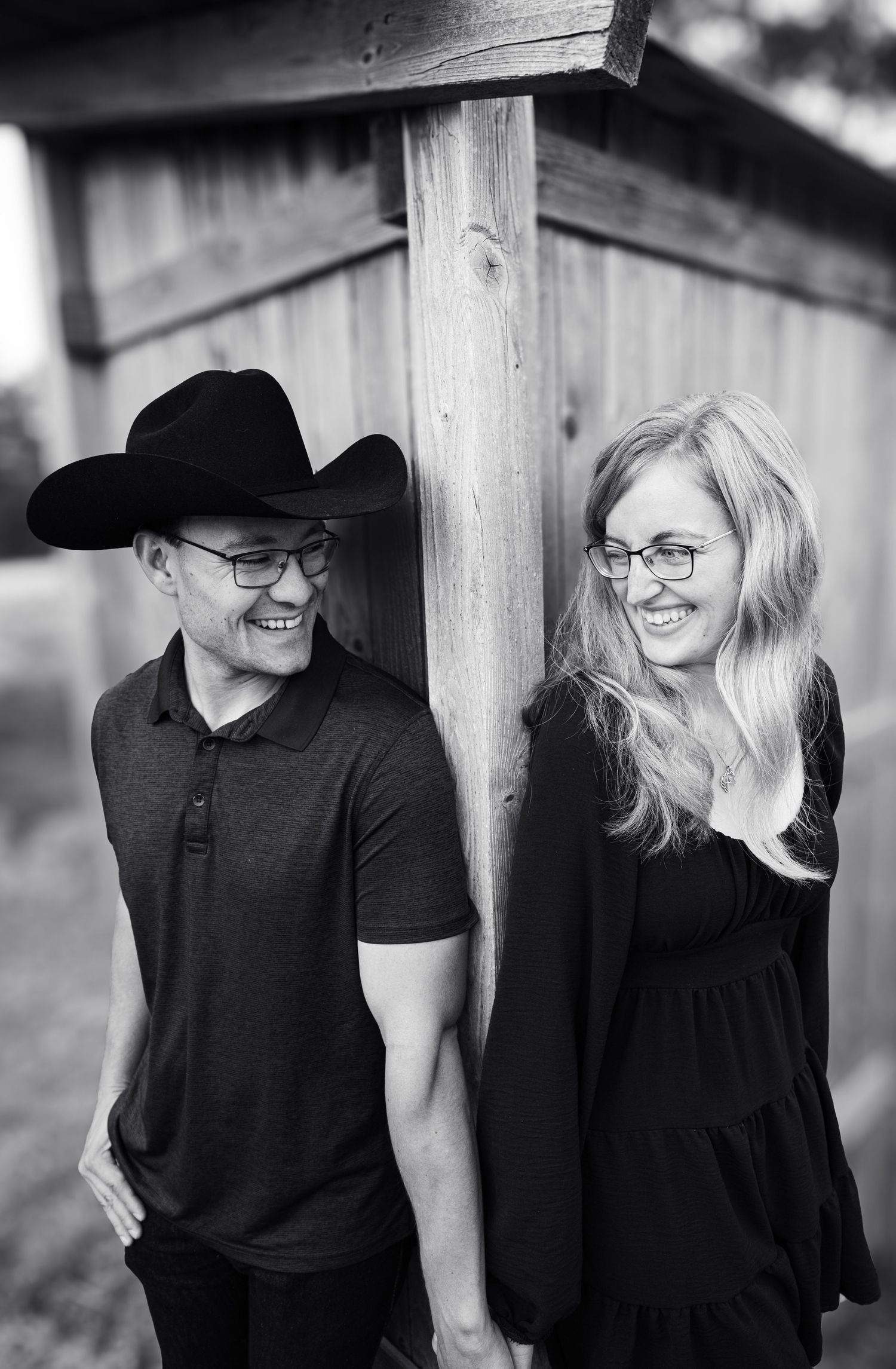 Smiling couple in glasses leaning against wooden post, man wearing black cowboy hat.