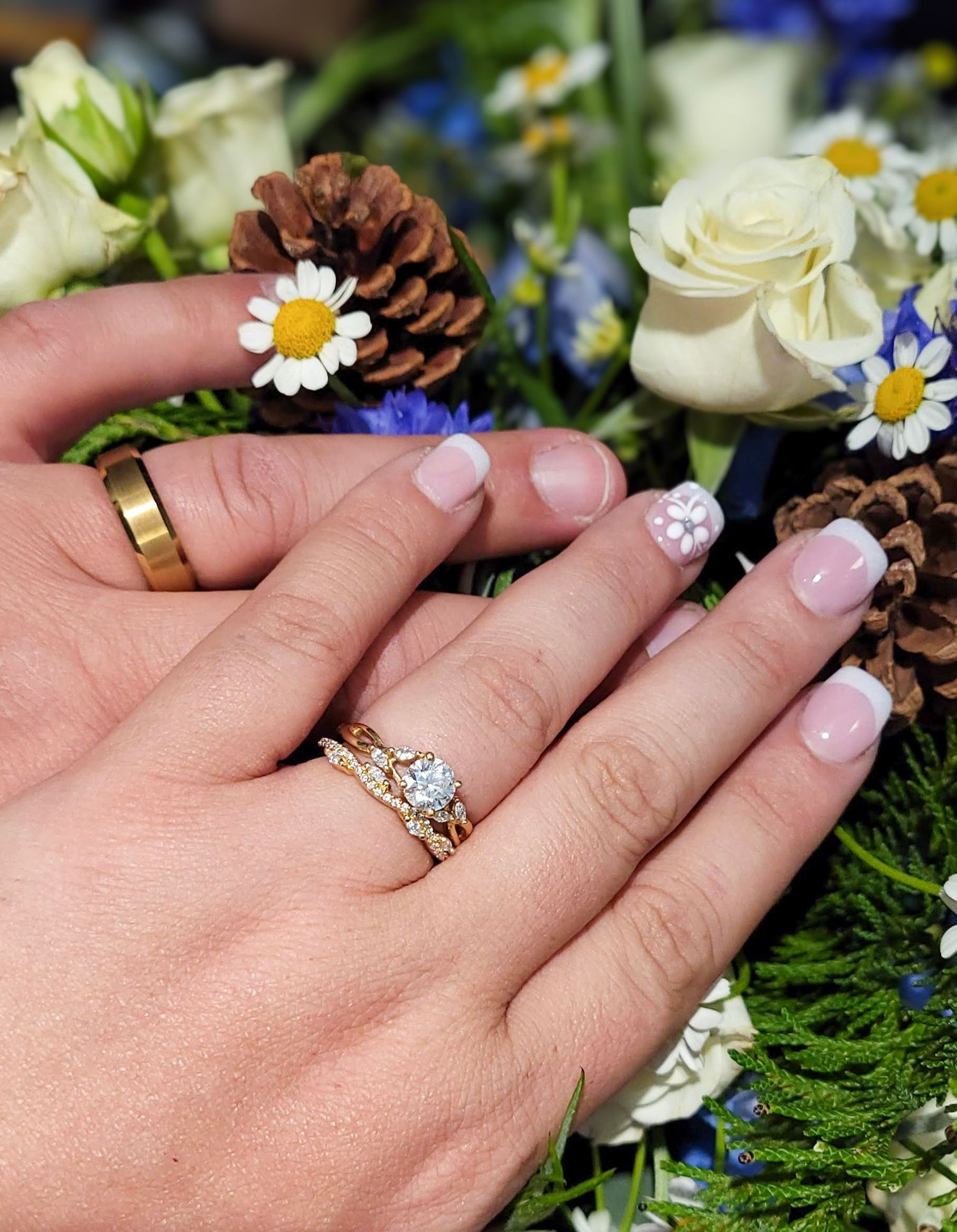 Close-up of wedding rings on bride and groom hands with floral bouquet featuring daisies and pinecones.