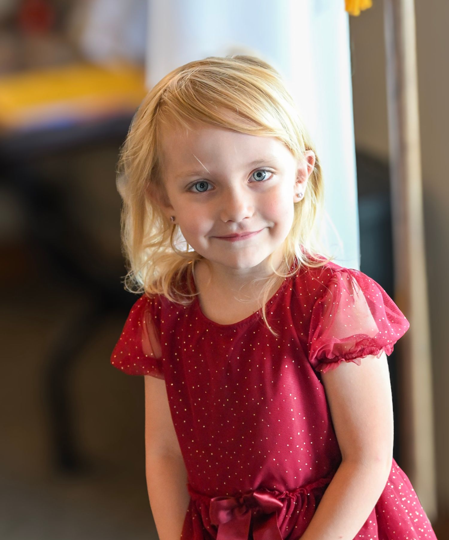 Smiling young blonde girl in a red sparkle dress with bow, wearing pearl earrings, posed indoors.