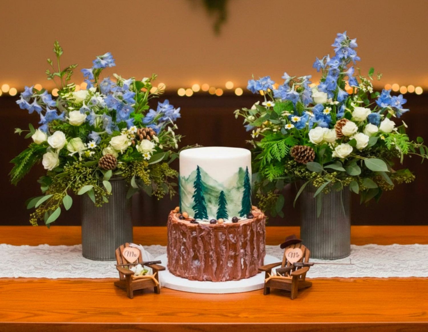 Elegant woodland wedding cake with painted pine trees flanked by blue and white floral arrangements on a wood table.