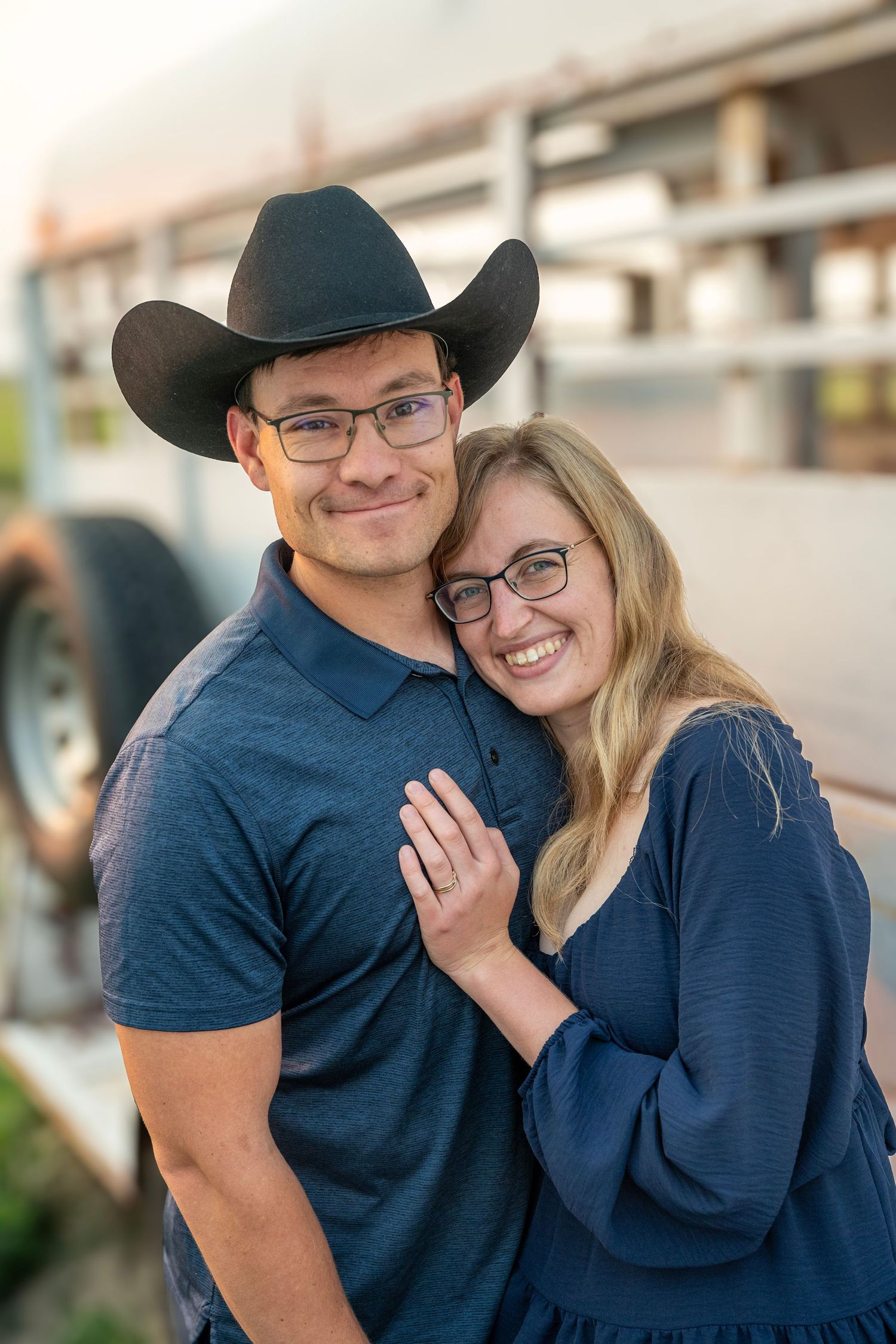 Smiling couple in matching blue outfits embracing near a horse trailer during engagement photos.