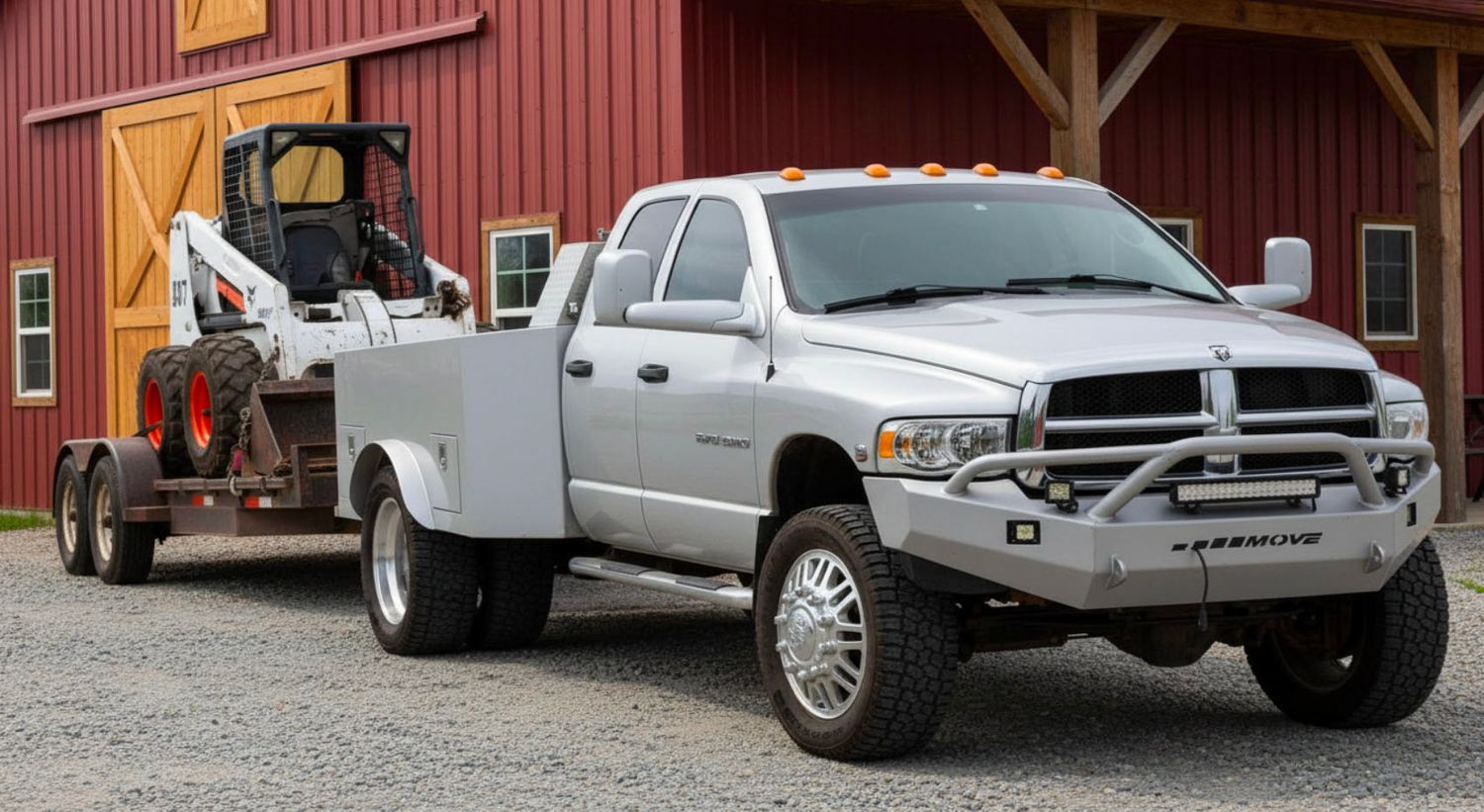 Silver Dodge Ram heavy duty pickup truck towing a flatbed trailer with a skid steer loader near a red barn.