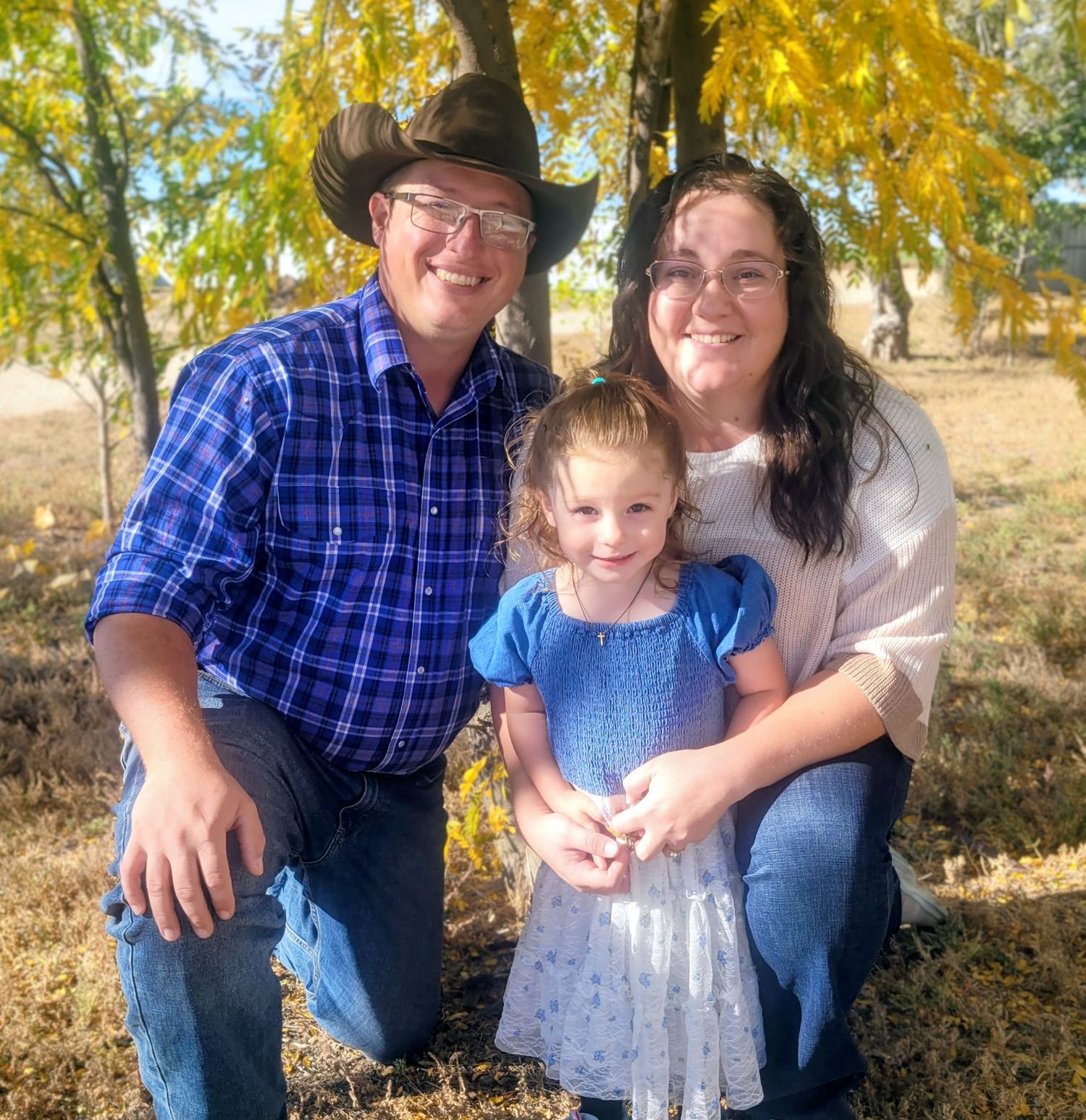 Happy family with father in cowboy hat, mother, and young daughter posing under autumn tree.