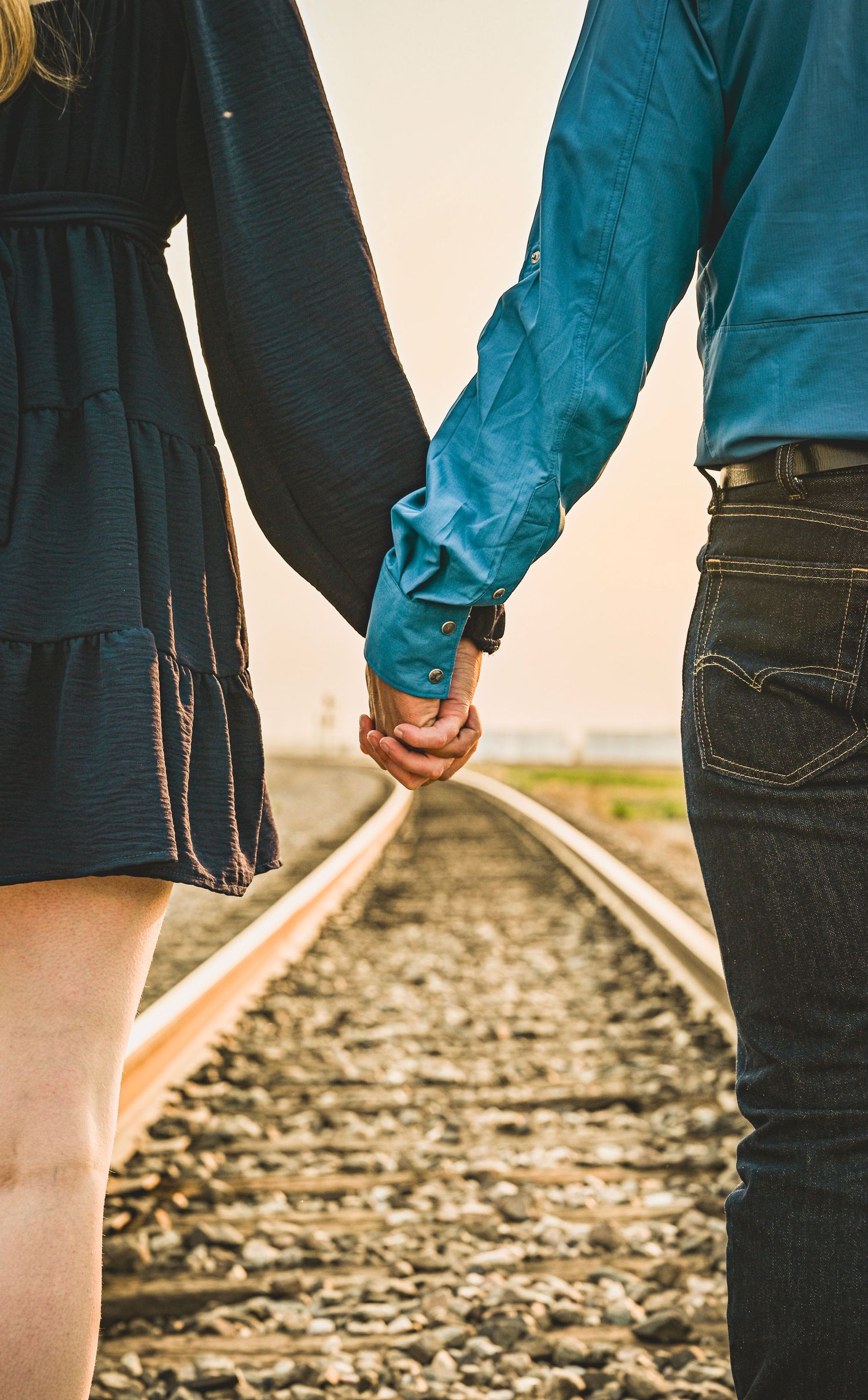 Couple holding hands while standing on railroad tracks, viewed from behind.