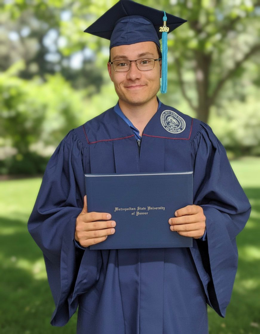 Graduate in navy cap and gown holding Metropolitan State University of Denver diploma outdoors.