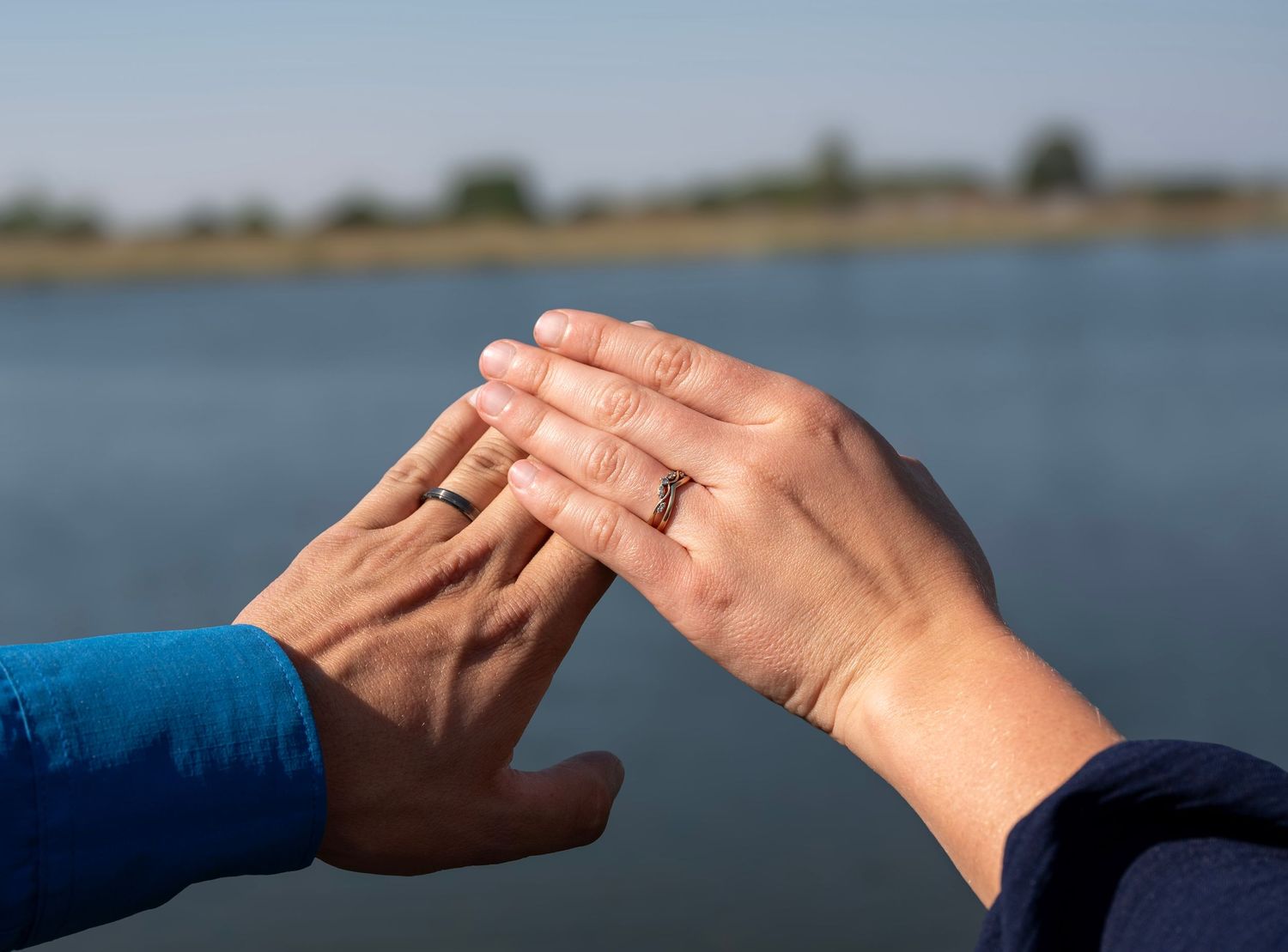 Newlywed couple touching hands showing wedding rings outdoors near a serene lake backdrop.