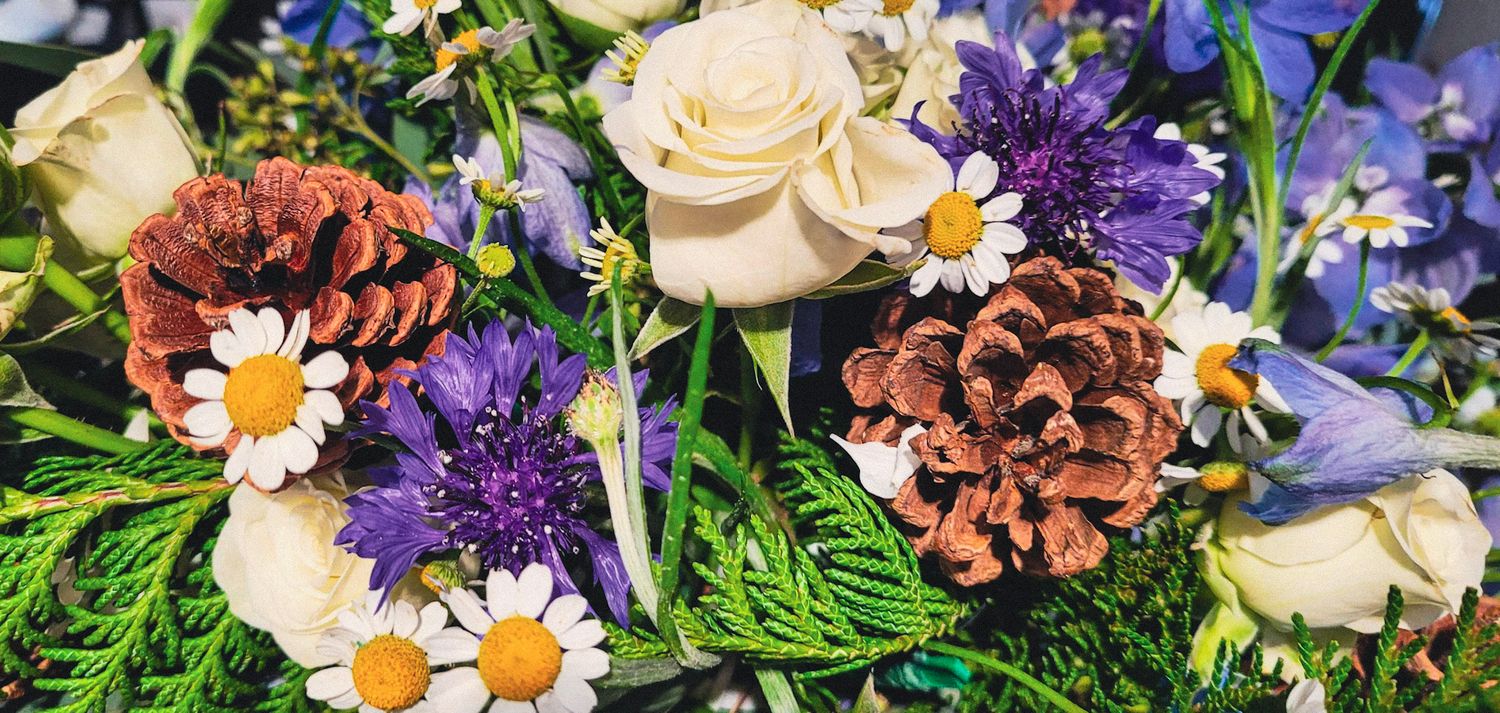 Rustic wedding bouquet with white roses, purple wildflowers, daisies, and decorative pinecones.