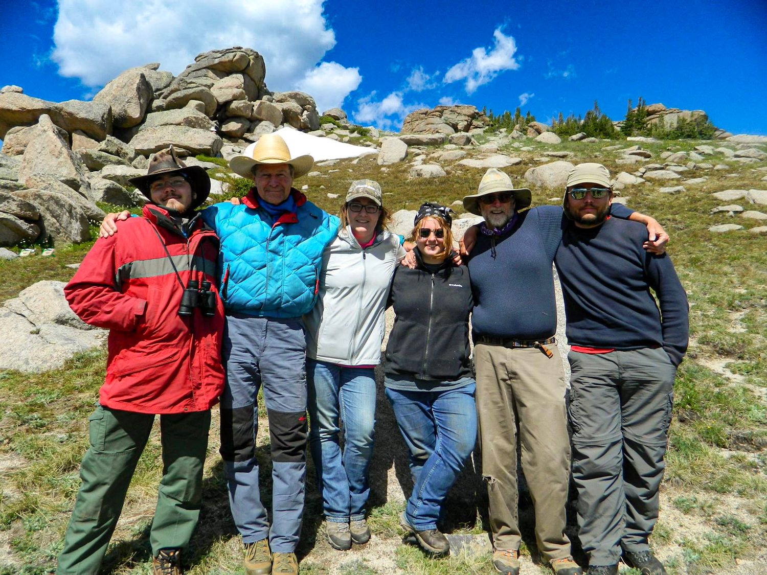Group of six hikers posing together in rocky mountain meadow on sunny day.