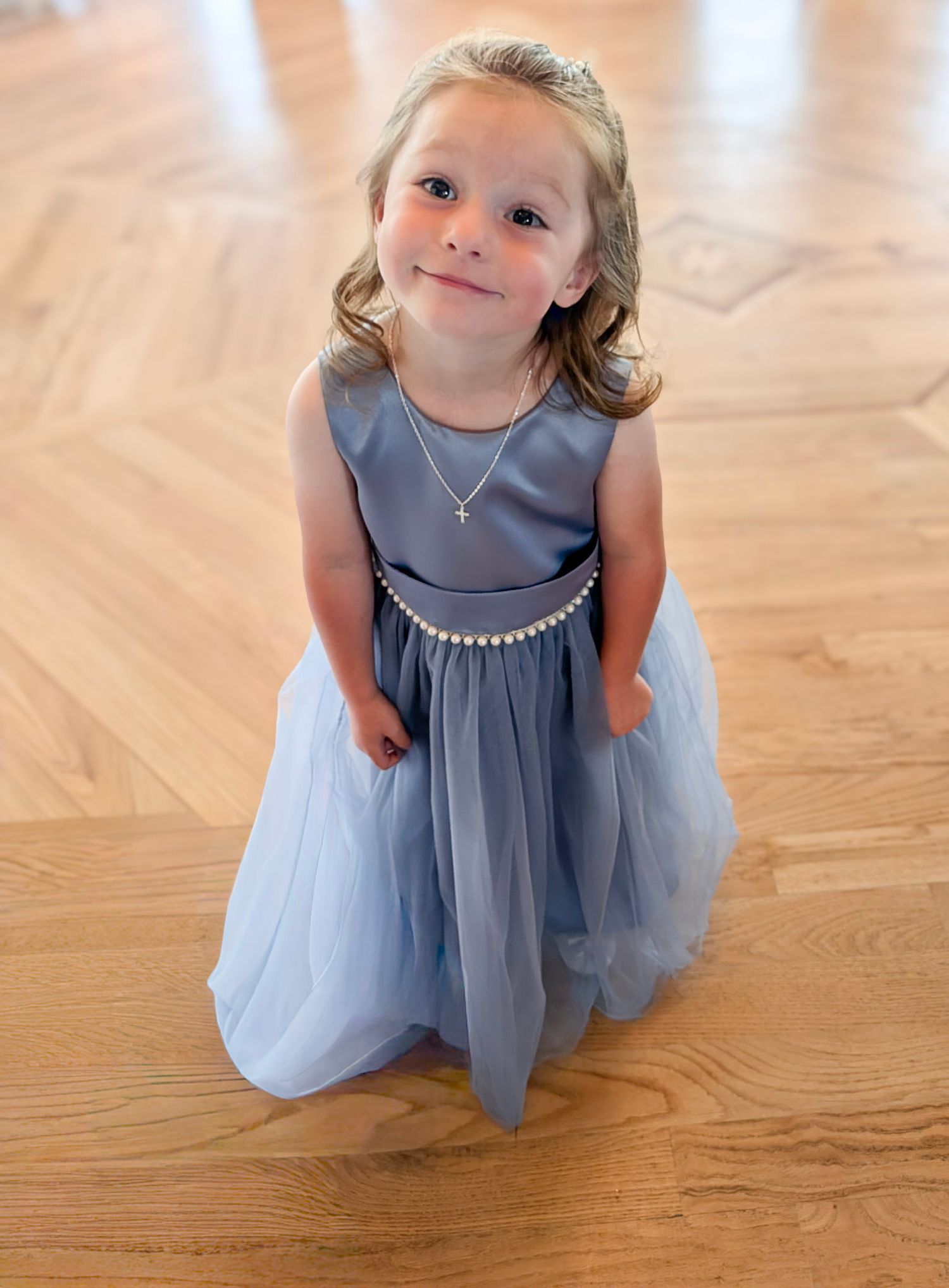 A young girl in a dusty blue tulle flower girl dress with pearl belt and necklace stands on hardwood floor.