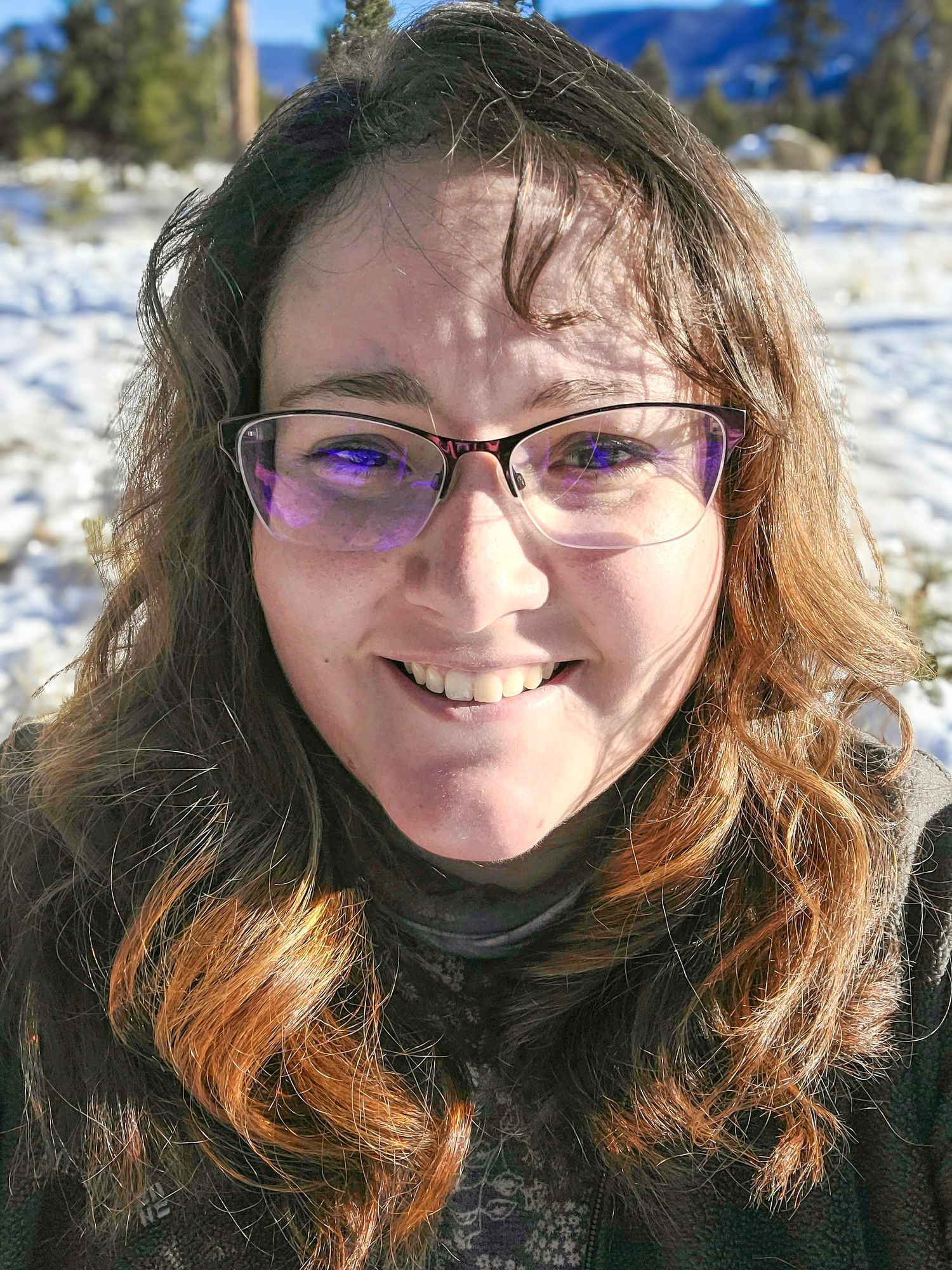 Smiling woman with glasses and wavy brown hair poses outdoors in a snowy winter mountain landscape.