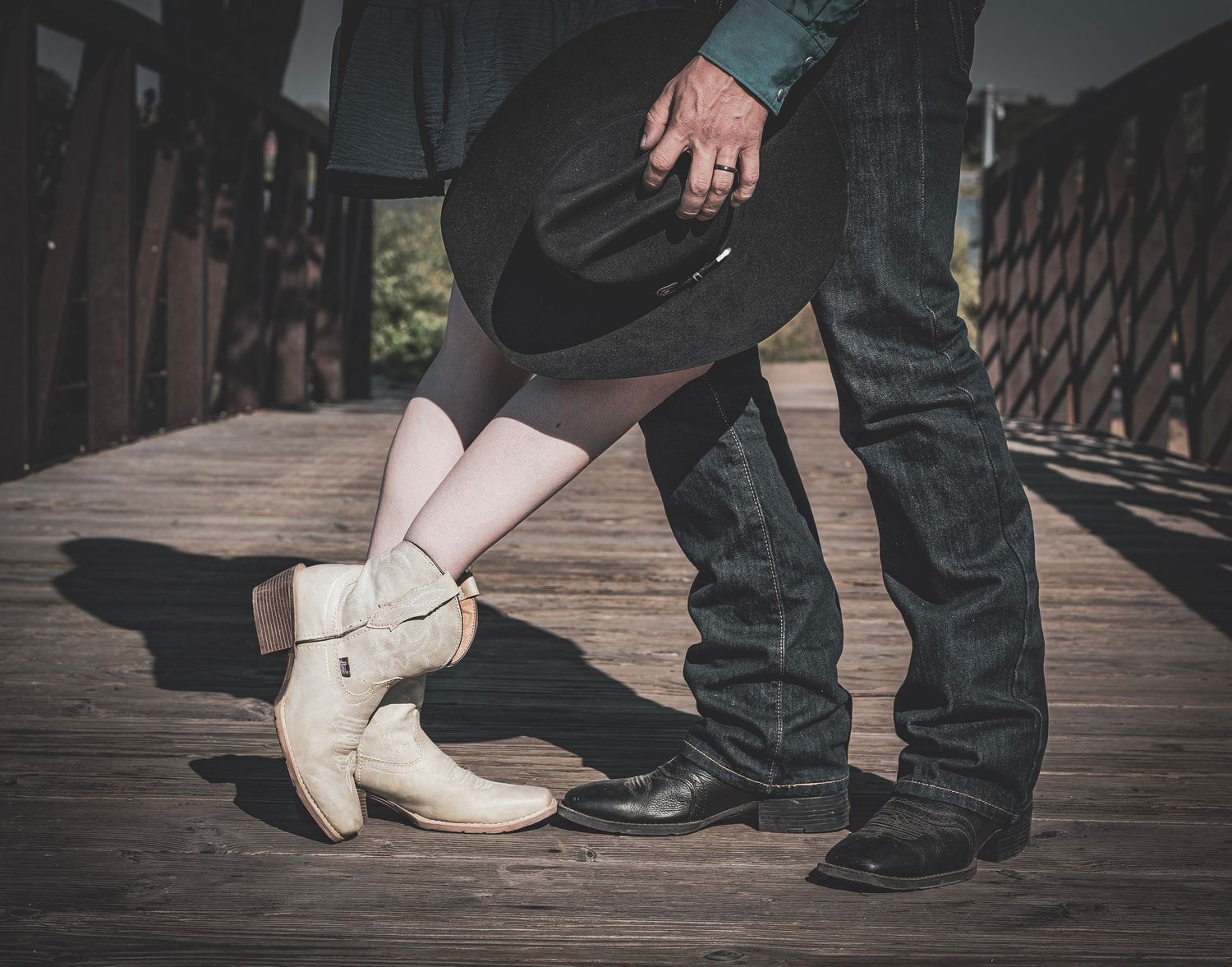 Couple in cowboy boots and hat holding hands on rustic wooden bridge during engagement photo.