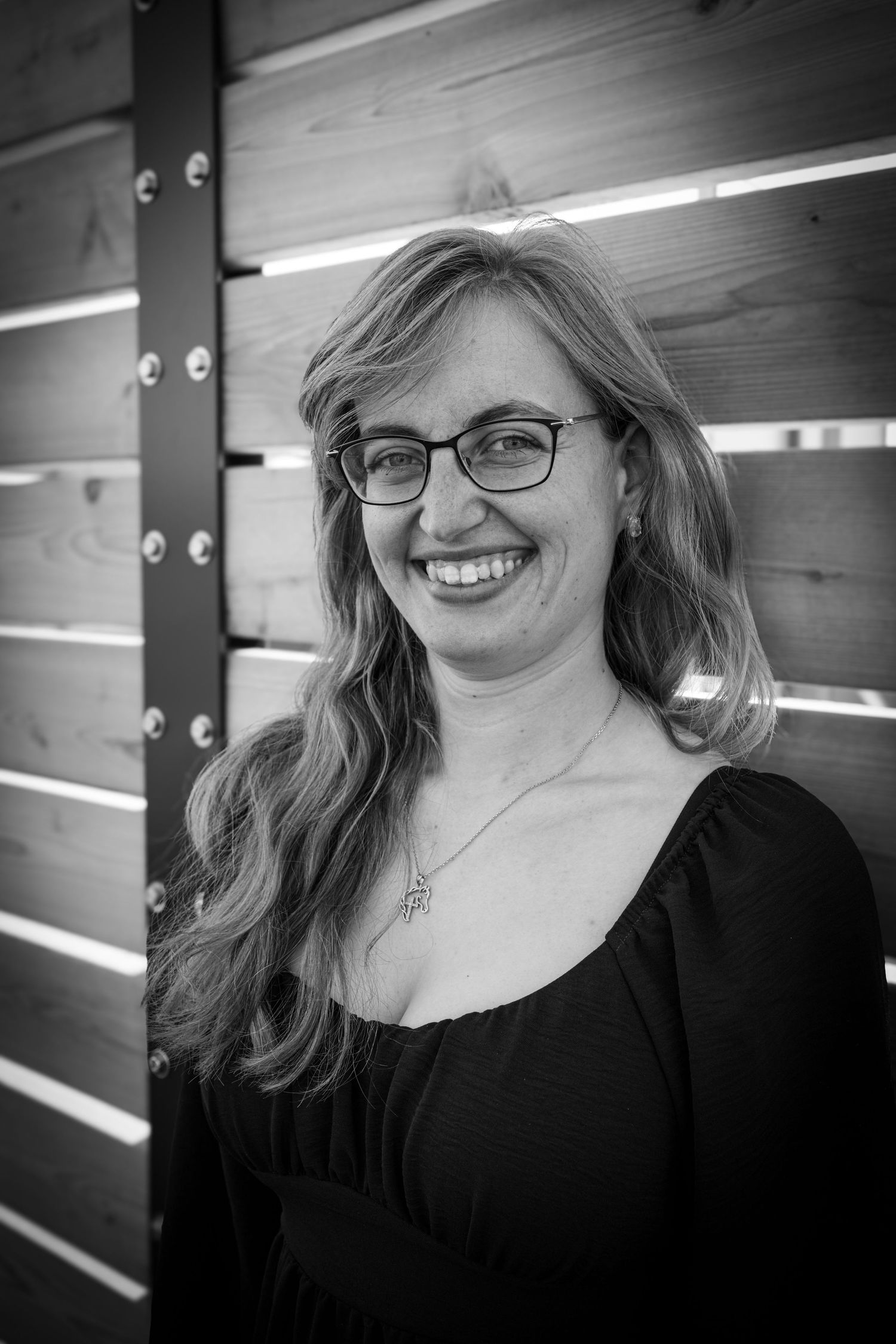 Black and white portrait of smiling woman with glasses leaning against wooden slat wall.