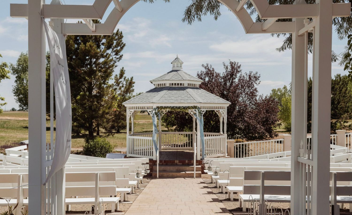 White gazebo wedding ceremony setup with rows of white chairs on a brick path, surrounded by lush greenery.