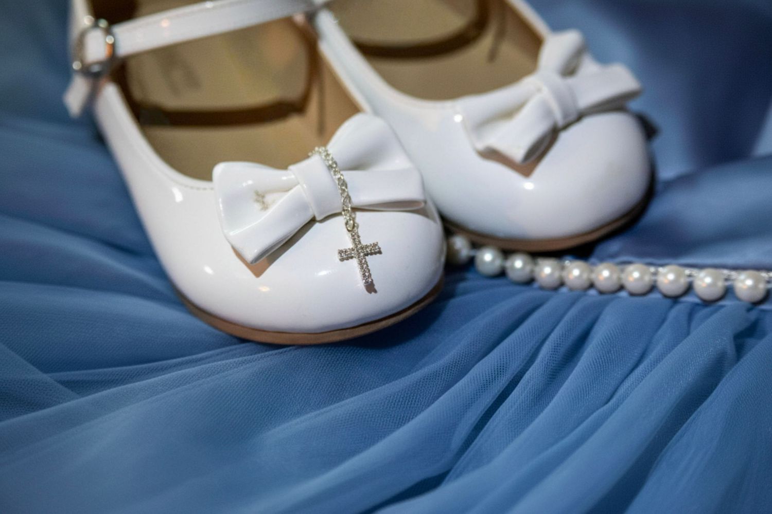 White patent leather flower girl shoes with bow and cross charm beside pearl necklace on blue dress.