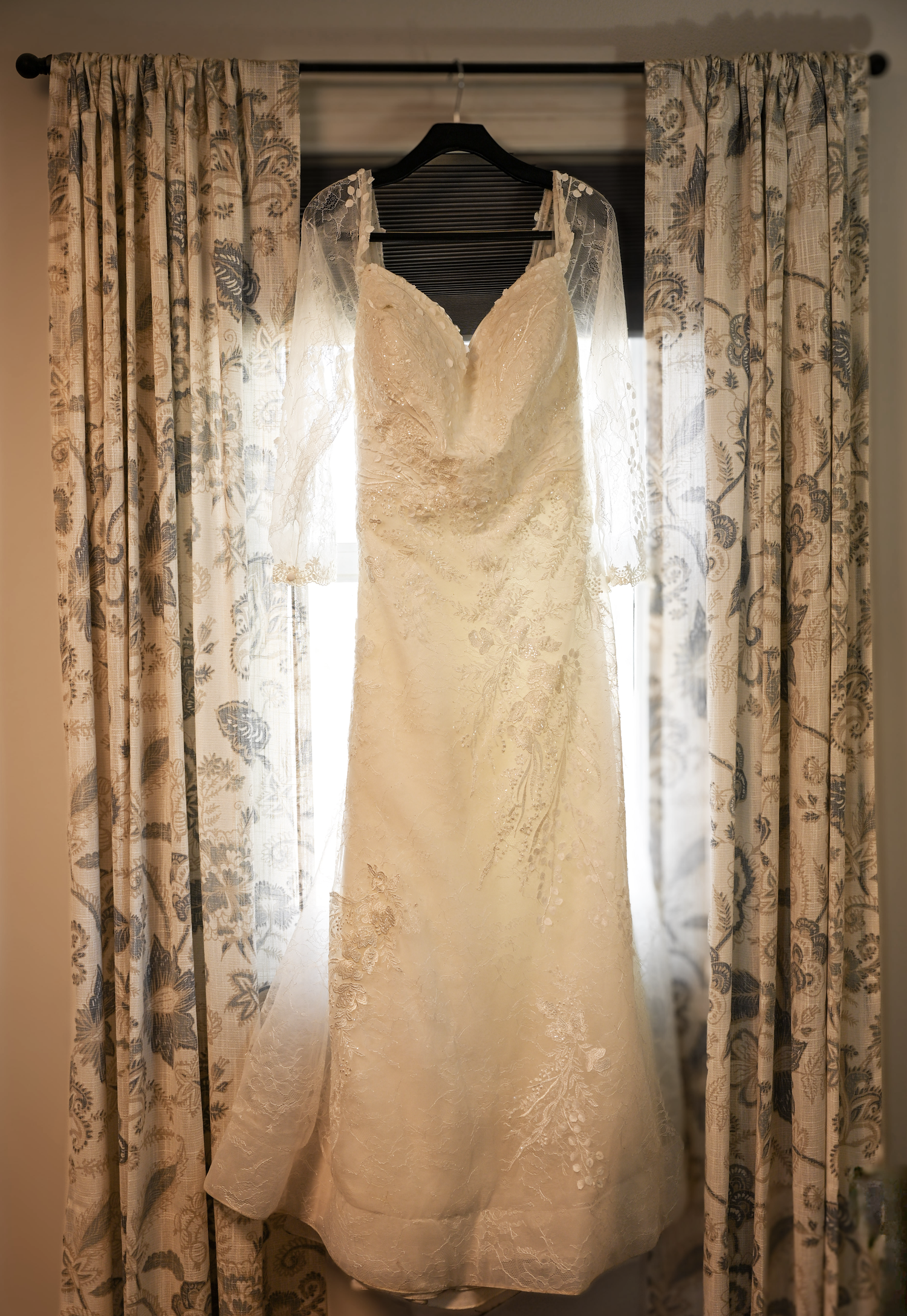 Elegant lace mermaid wedding dress hanging in window with floral curtains and soft natural light.