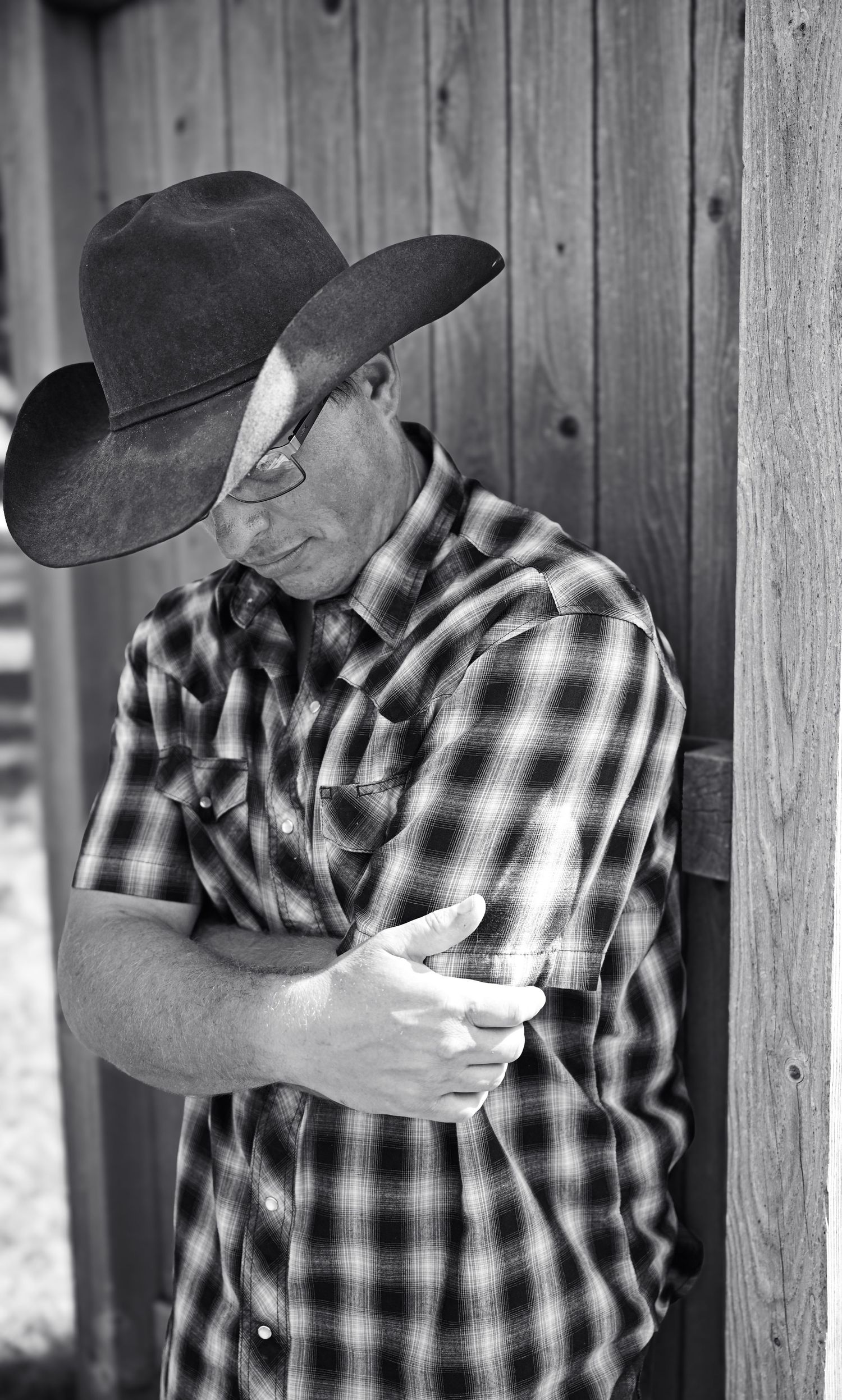 Black and white portrait of a man in cowboy hat and plaid shirt leaning against a barn.