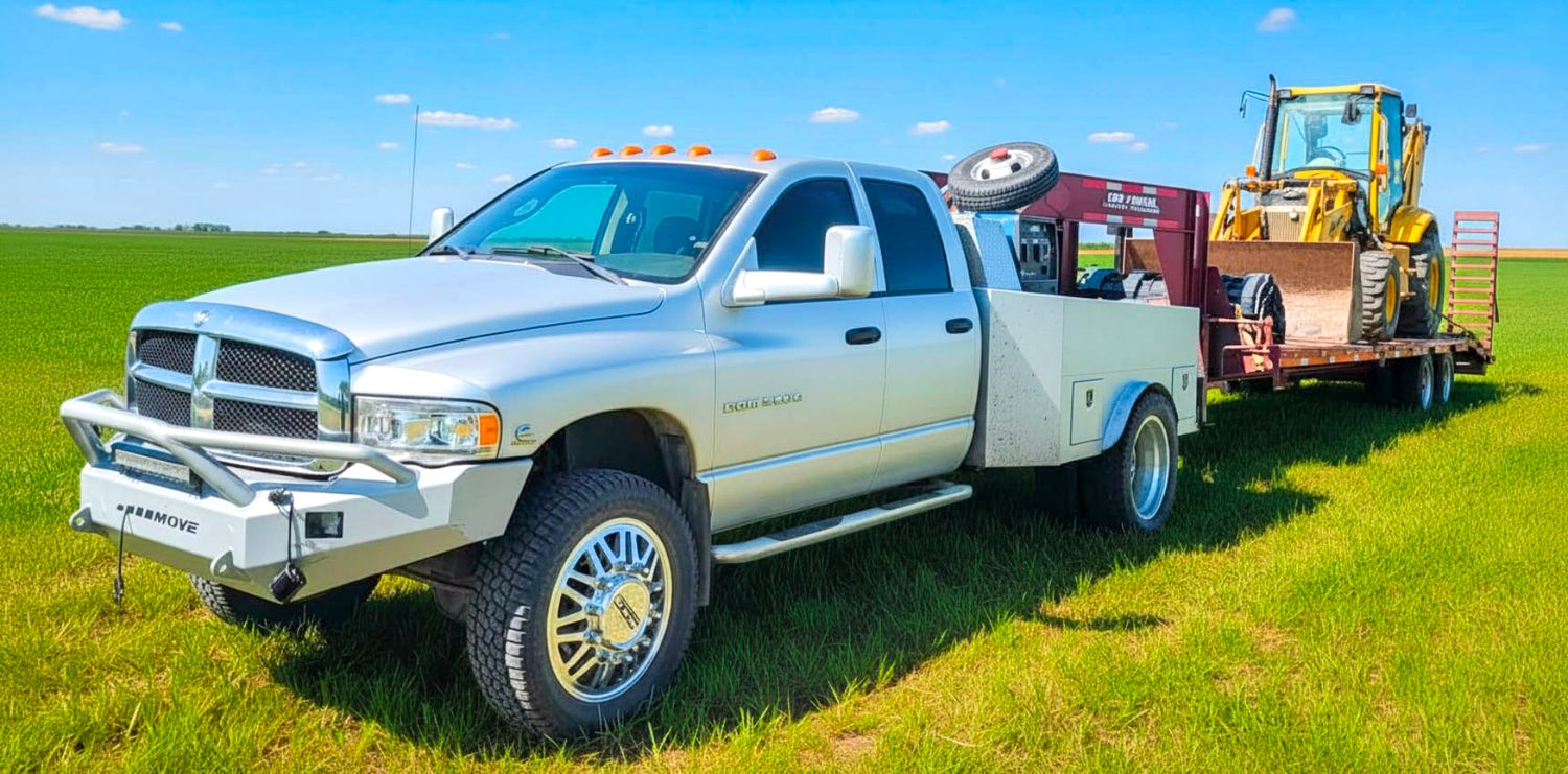 Silver Dodge Ram 4x4 pickup truck towing farm equipment on a flatbed trailer in a green field.