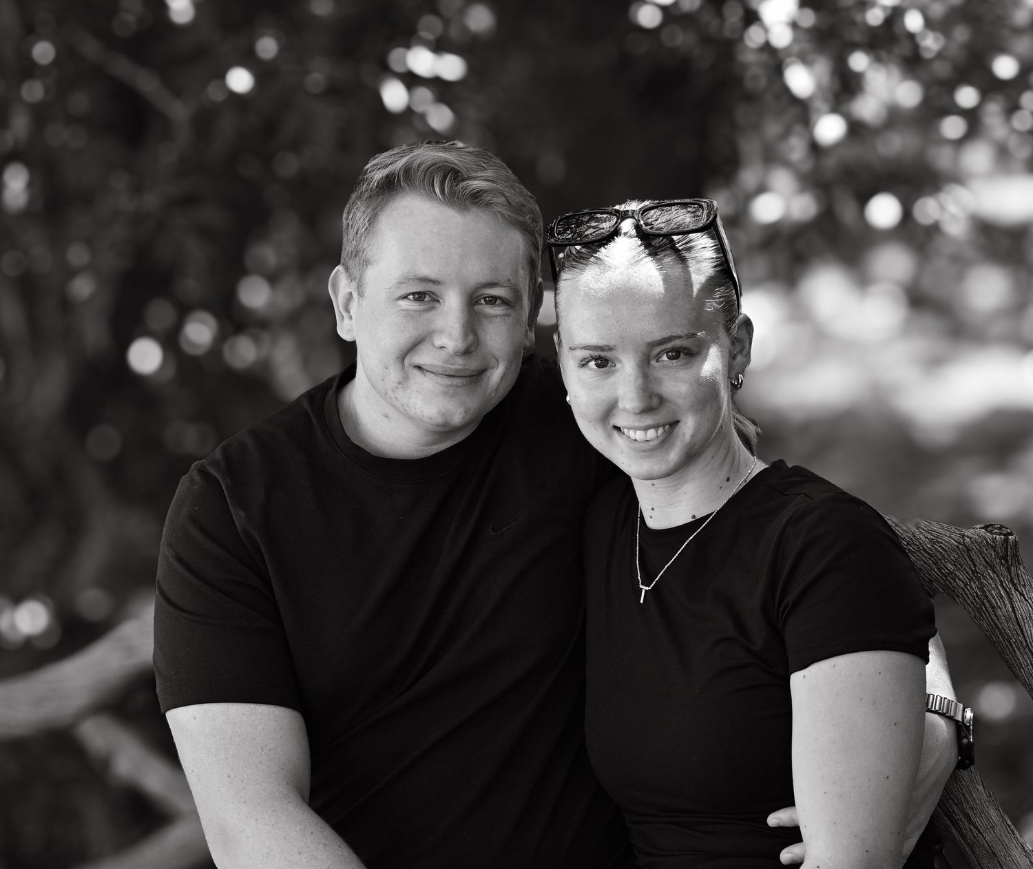 Black and white portrait of a smiling couple in black t-shirts posing outdoors with blurred trees behind them.