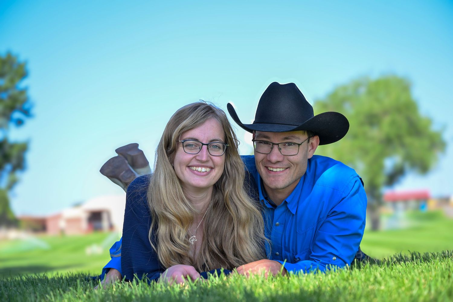 Happy couple lying on green grass smiling, man wearing black cowboy hat on sunny day.