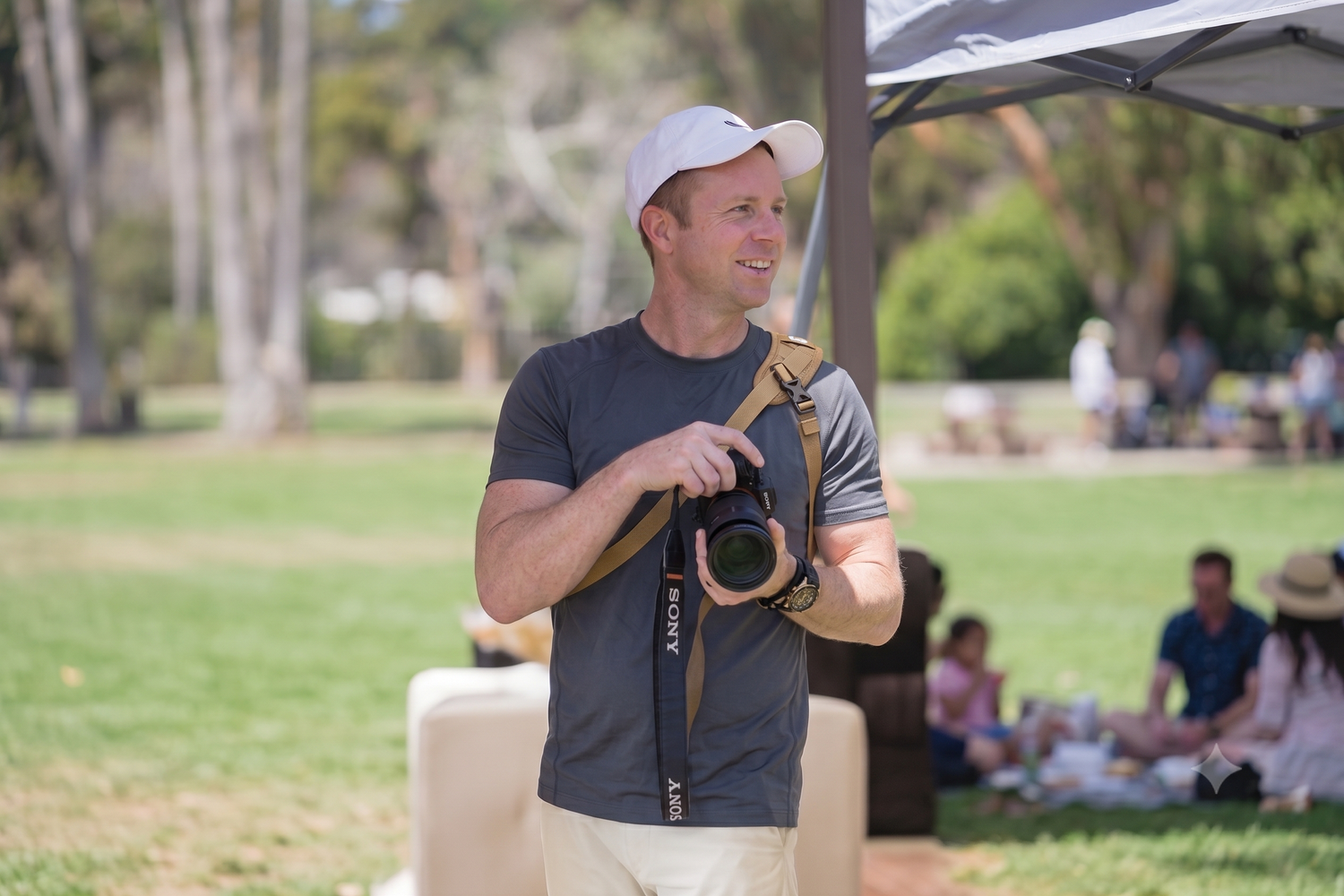 Man in gray shirt holding a Sony camera outdoors in a sunny park setting.