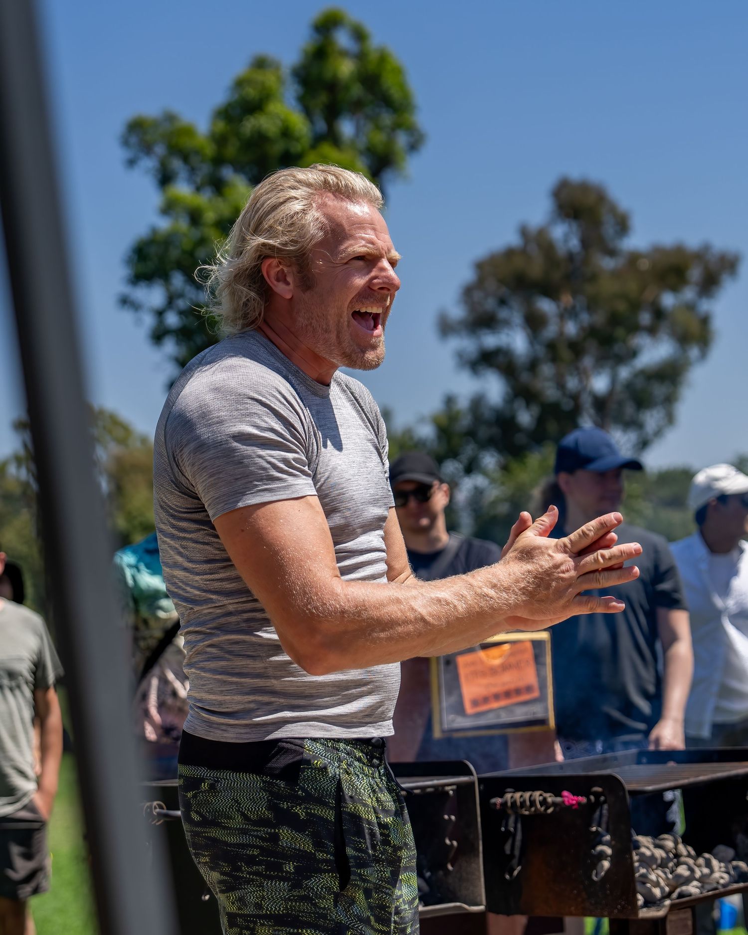 Energetic blond man clapping and laughing outdoors near a grill at a sunny park gathering.