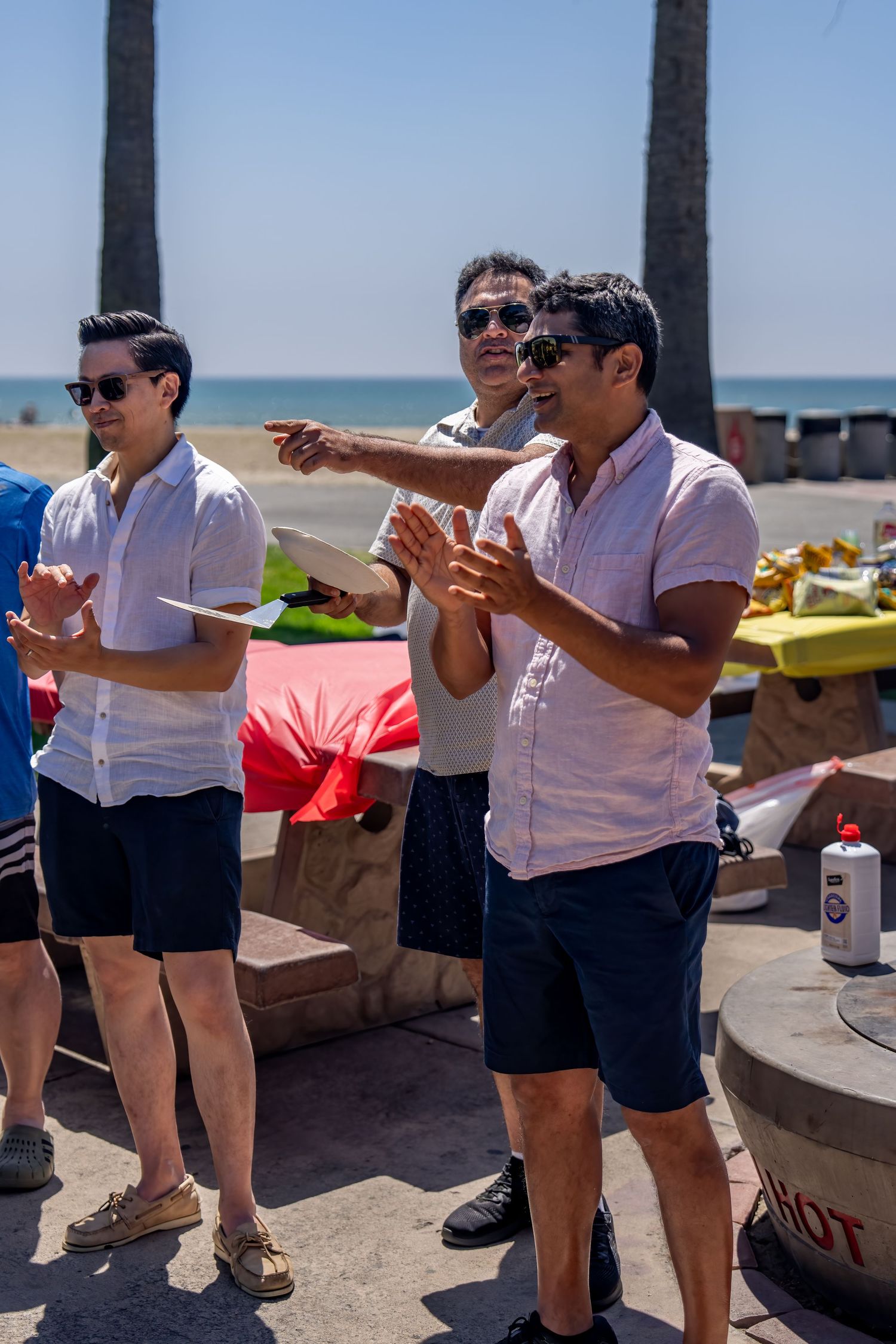 Three men in matching white shirts and navy shorts clapping at an outdoor beach barbecue event on a sunny day.