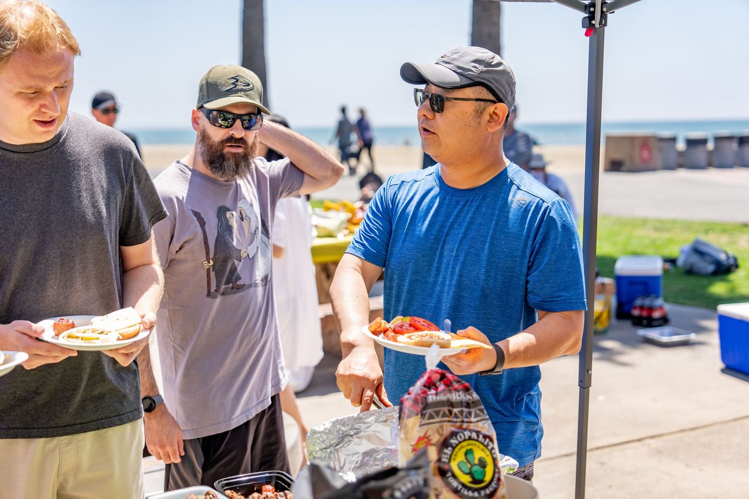 Men holding plates of food near a grill at a sunny outdoor beach barbecue event.