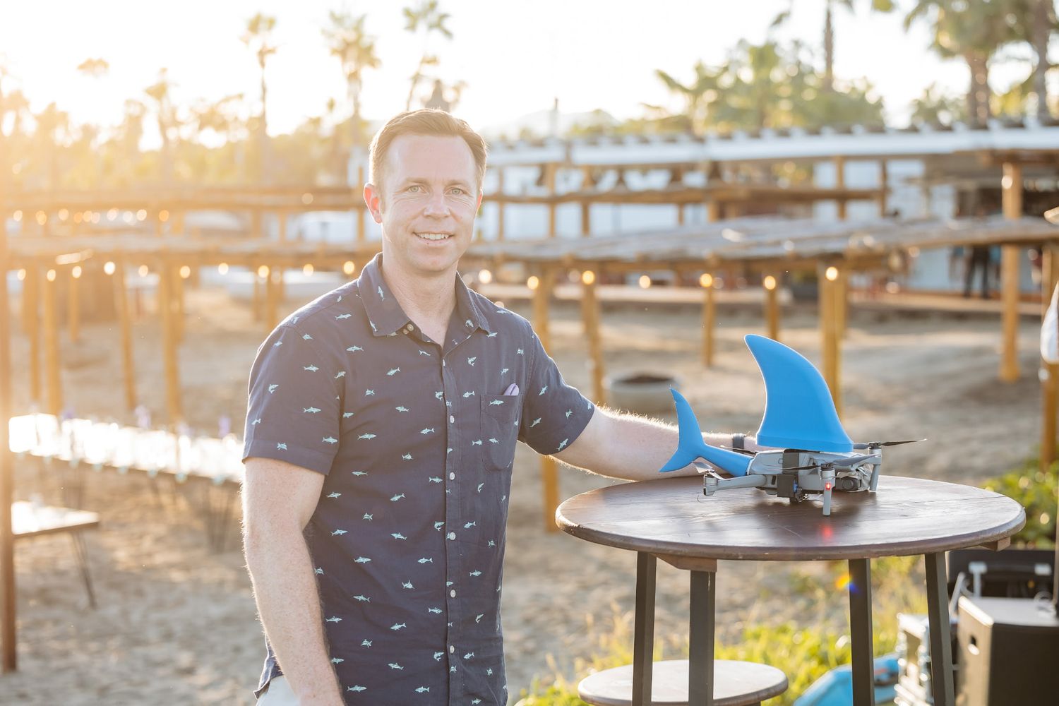 Man smiling next to a table displaying blue surfboard fins at a golden hour beach setting.
