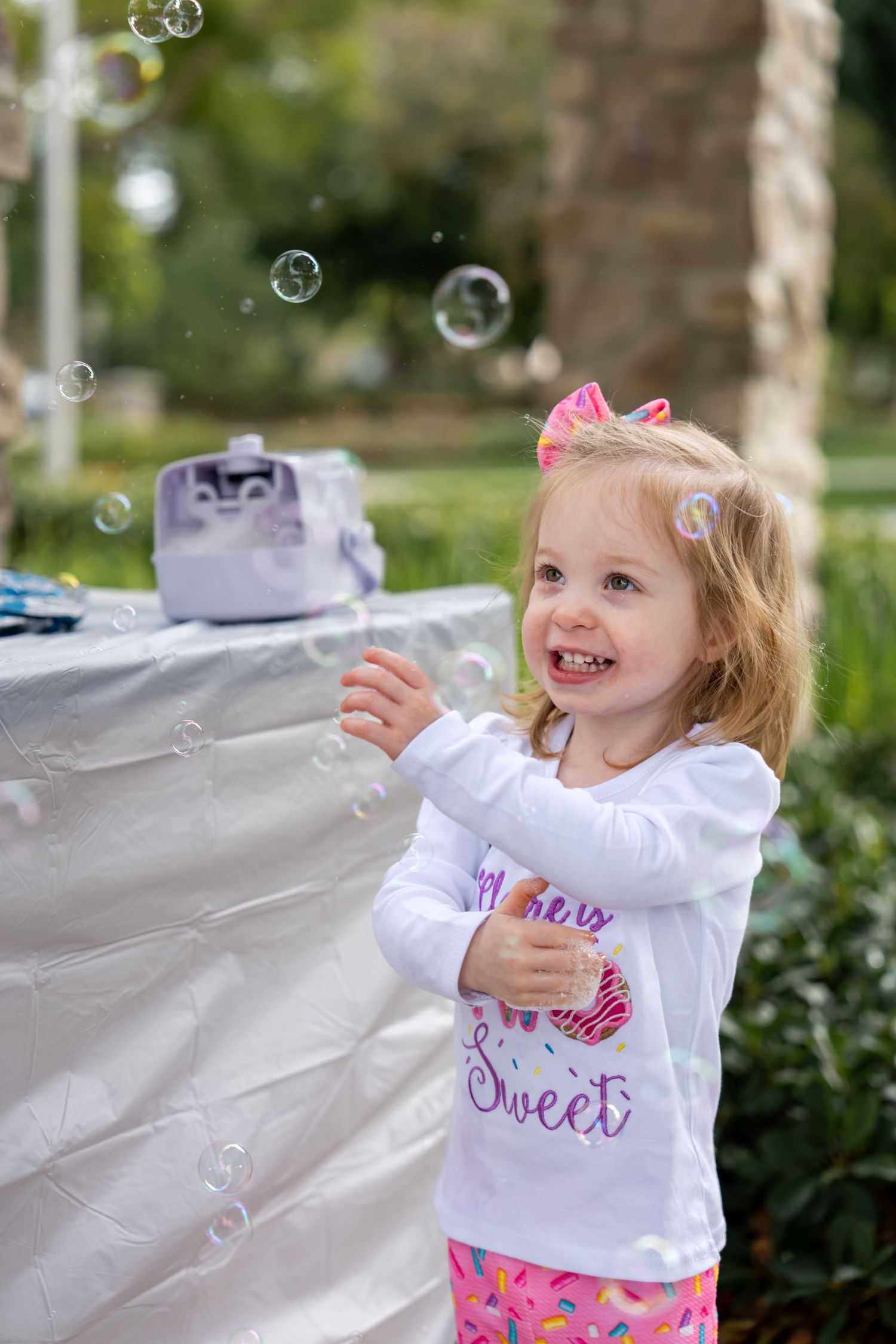 A happy toddler girl in a pink bow reaches for bubbles at an outdoor birthday party.