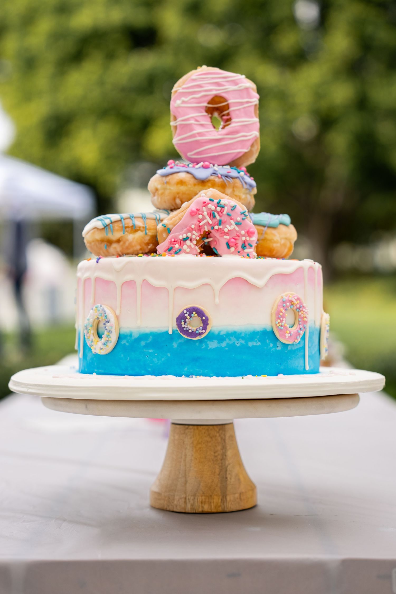 Colorful birthday cake decorated with donuts and pink frosting on a white cake stand outdoors.