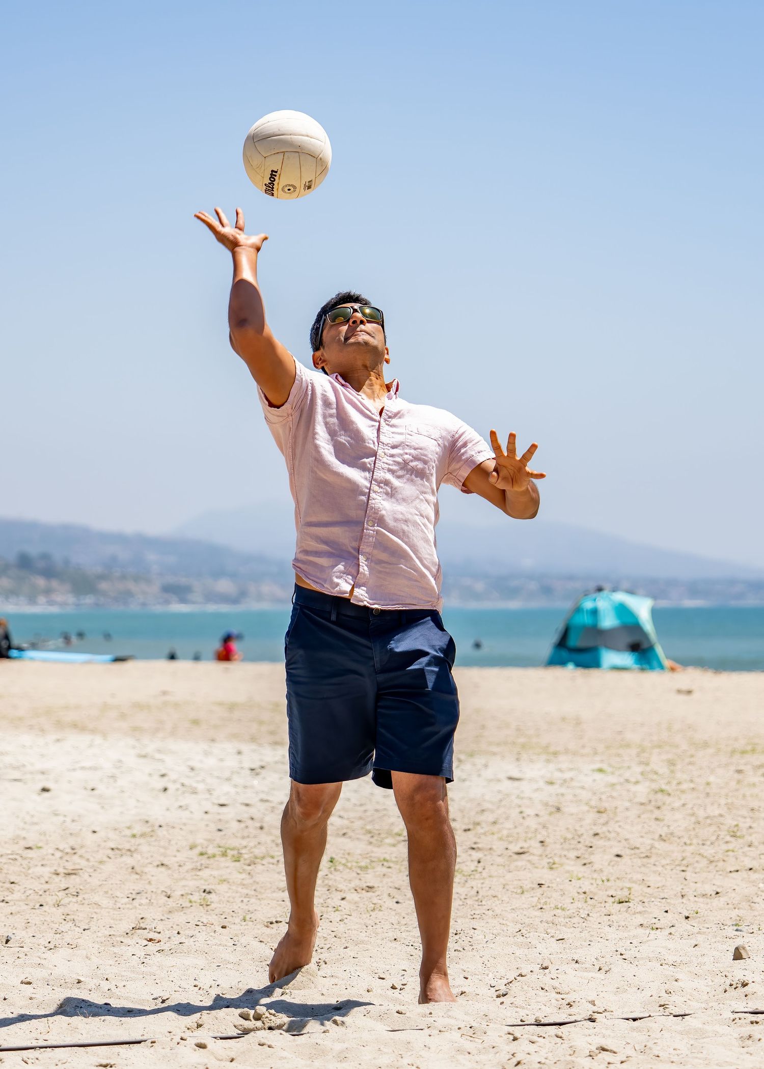 Man in pink shirt jumping to hit a white volleyball on a sunny beach with mountains behind him.