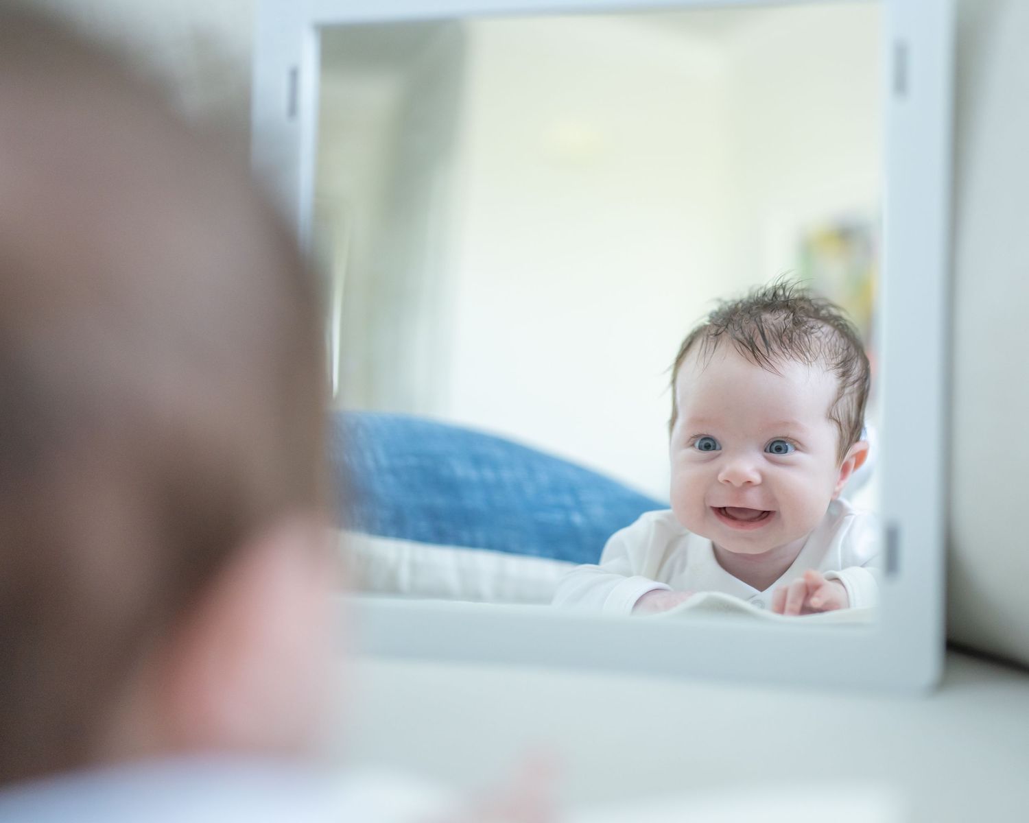 A smiling baby looks at their reflection in a small mirror while on tummy.
