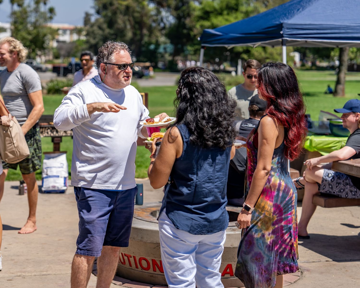 Man in white shirt gesturing while talking to two women at a sunny outdoor park gathering.