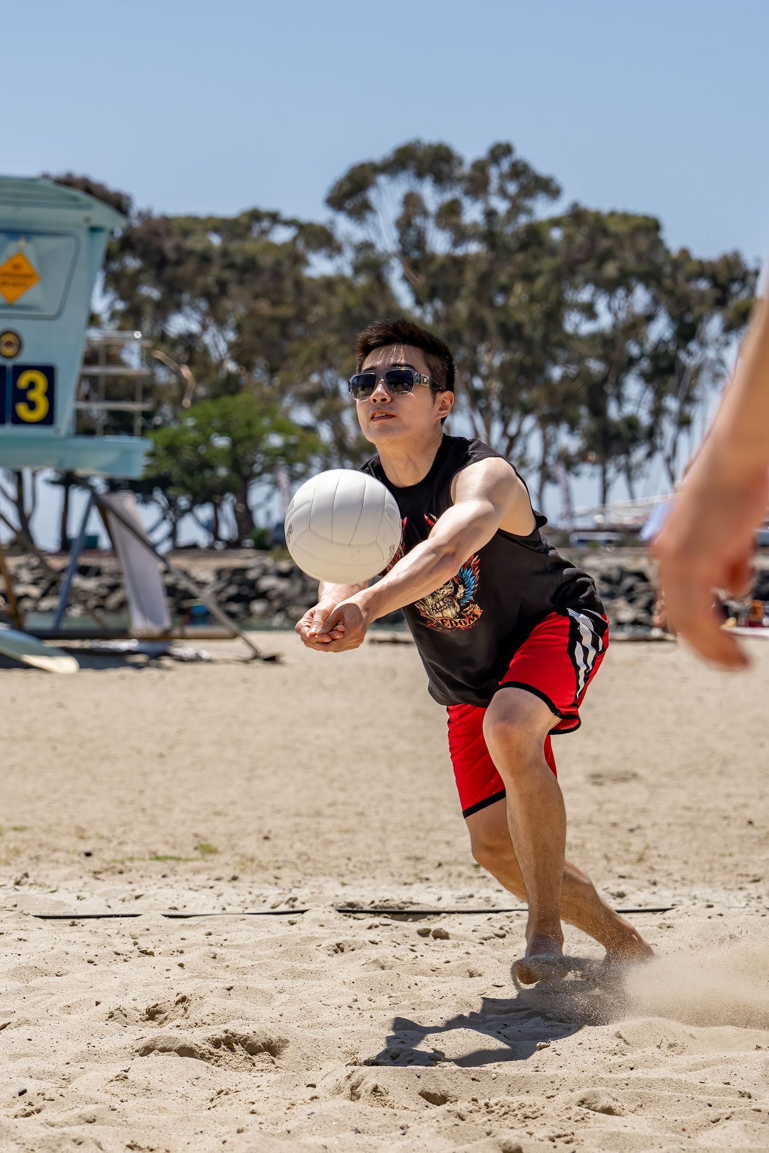 Athletic man in sunglasses bumps volleyball on sunny beach near lifeguard stand.