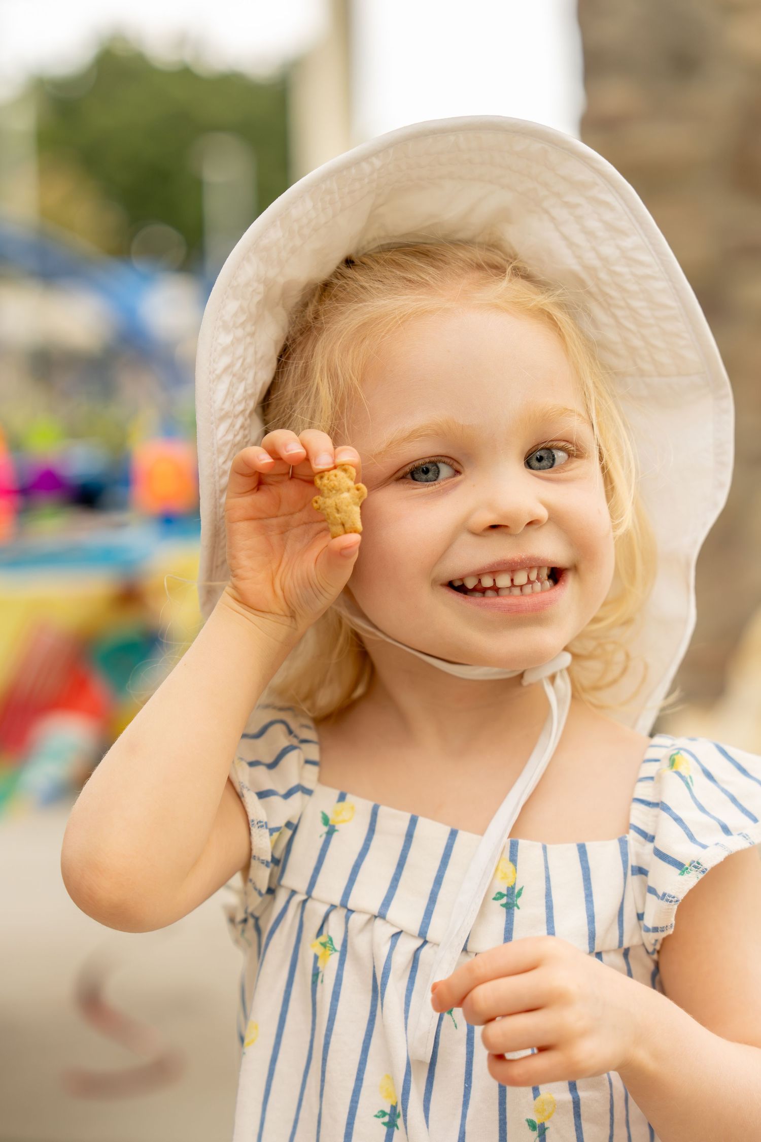 Cute blonde girl in a white hat smiling and holding a small snack at an outdoor fair.