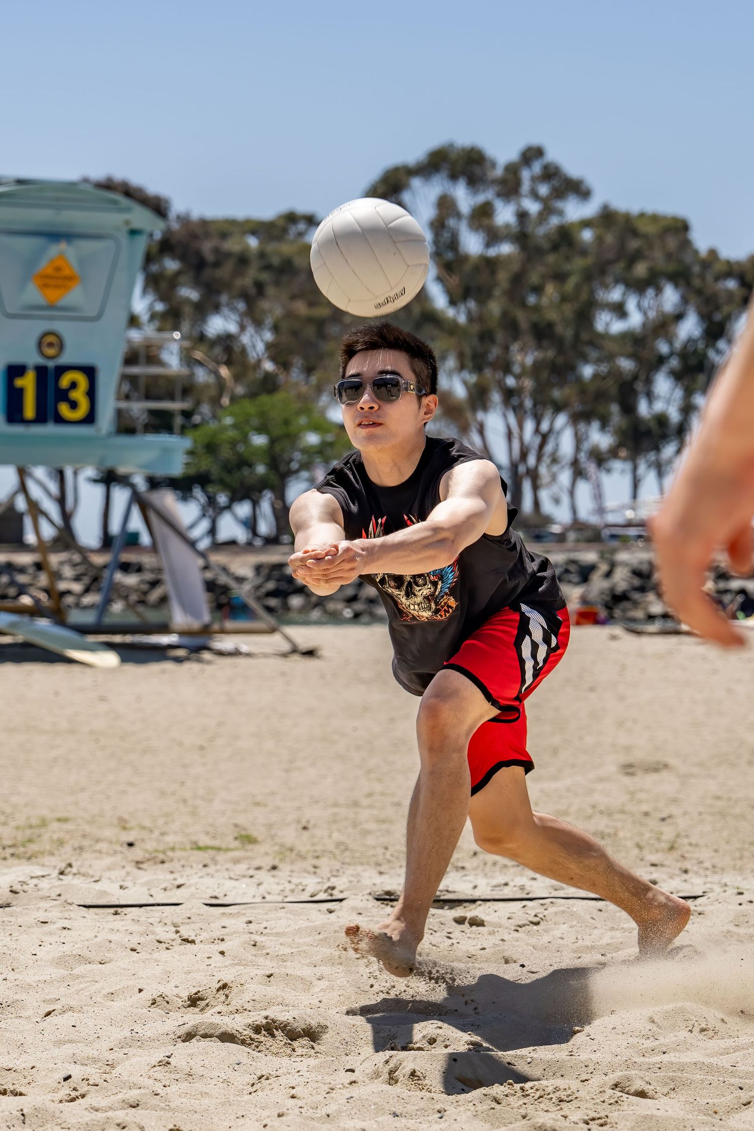 Young man in sunglasses digs volleyball on sandy beach by lifeguard tower.