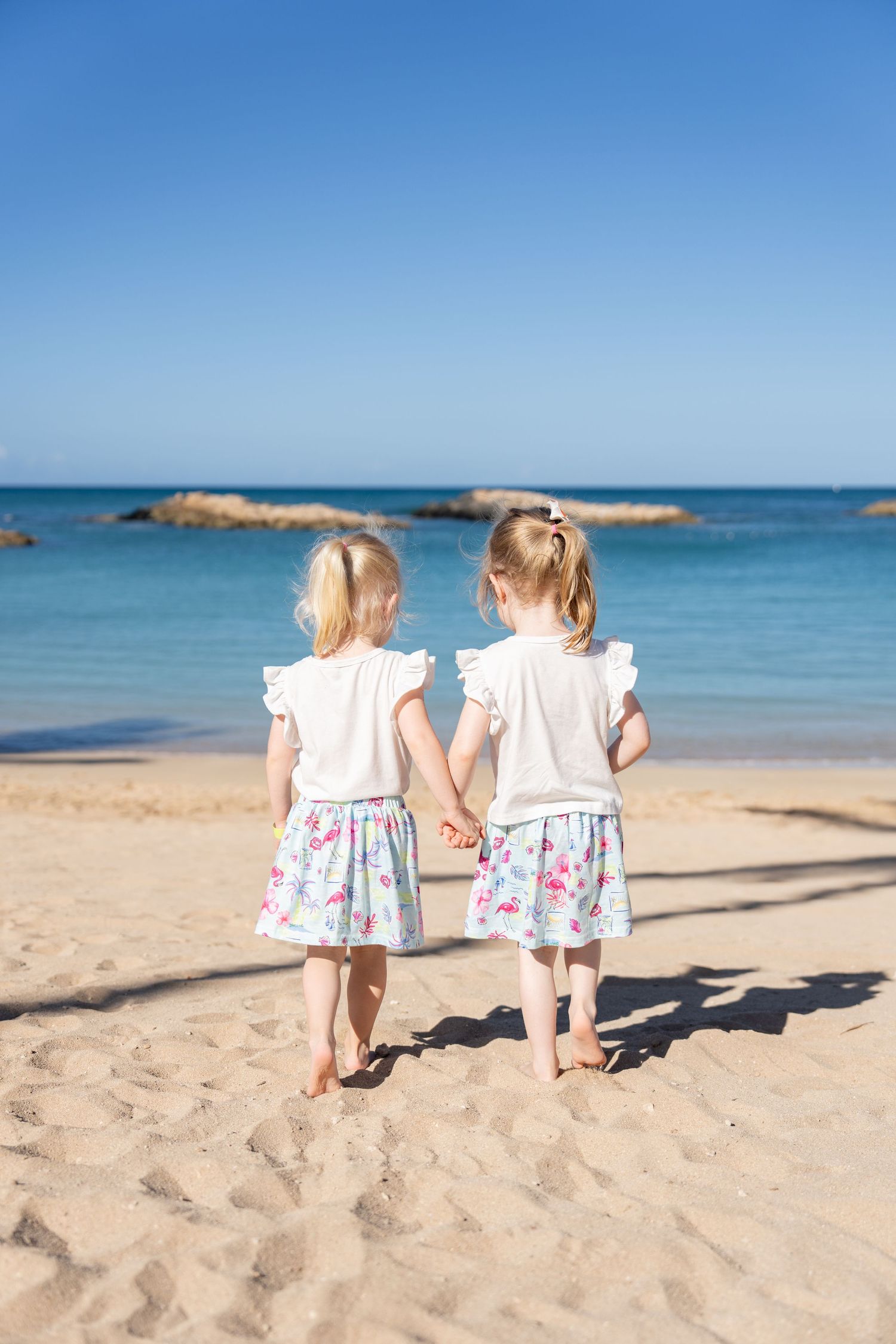 Two young blonde girls in matching floral skirts hold hands walking toward a calm turquoise beach.