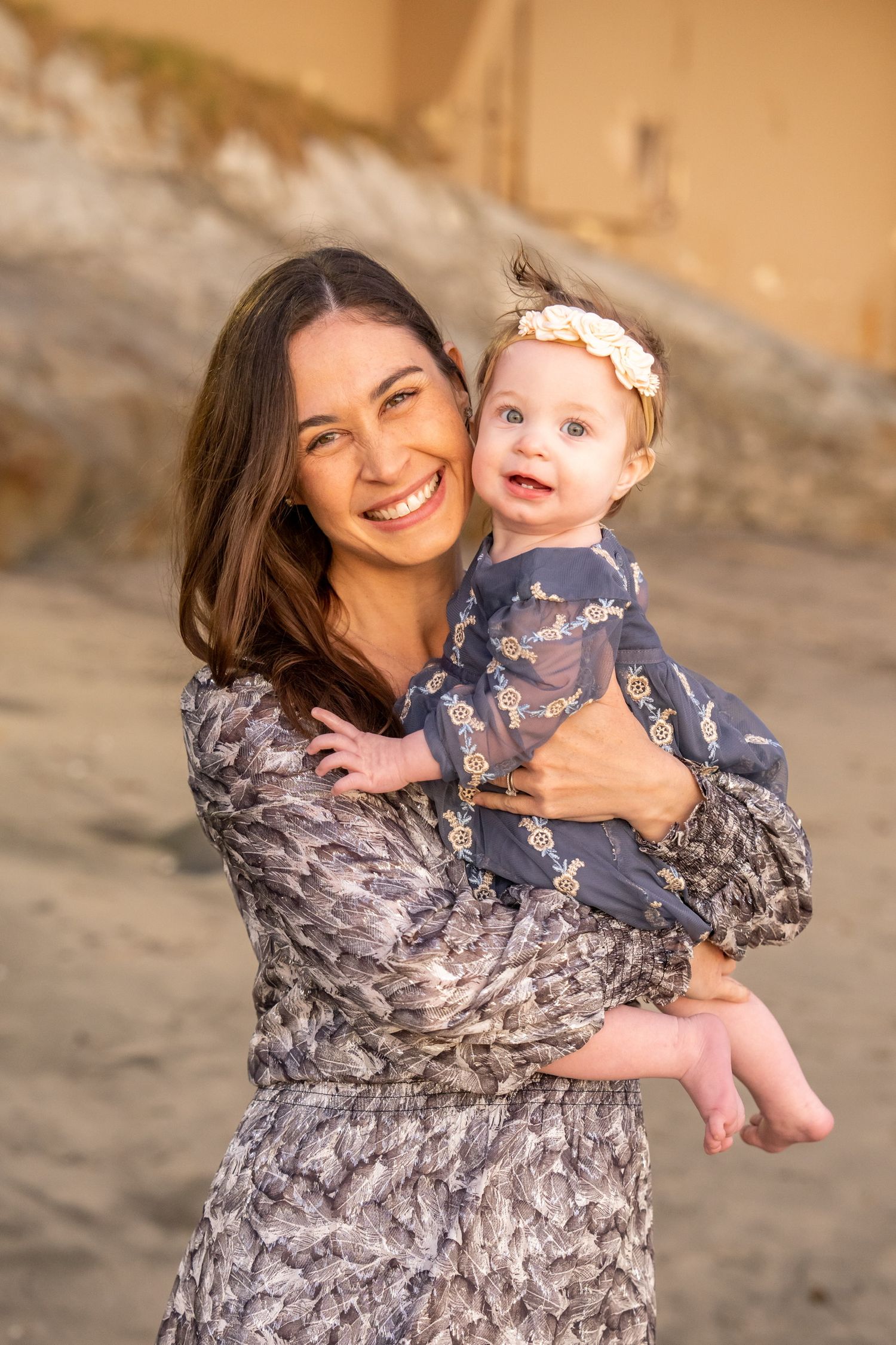 Smiling brunette mother holding happy baby girl with floral headband on sandy beach.