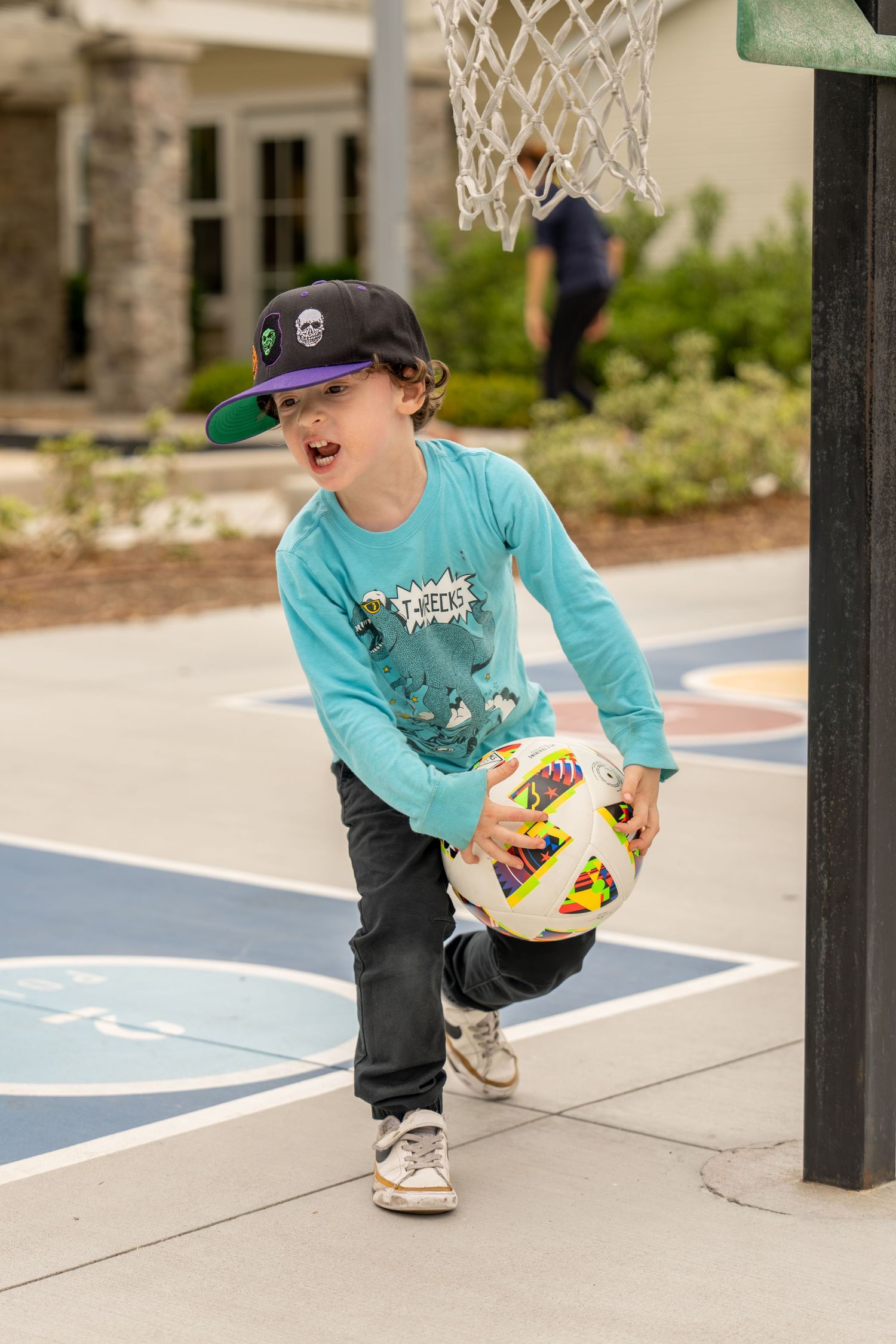 Young boy in teal shirt and cap dribbling a soccer ball on an outdoor basketball court.