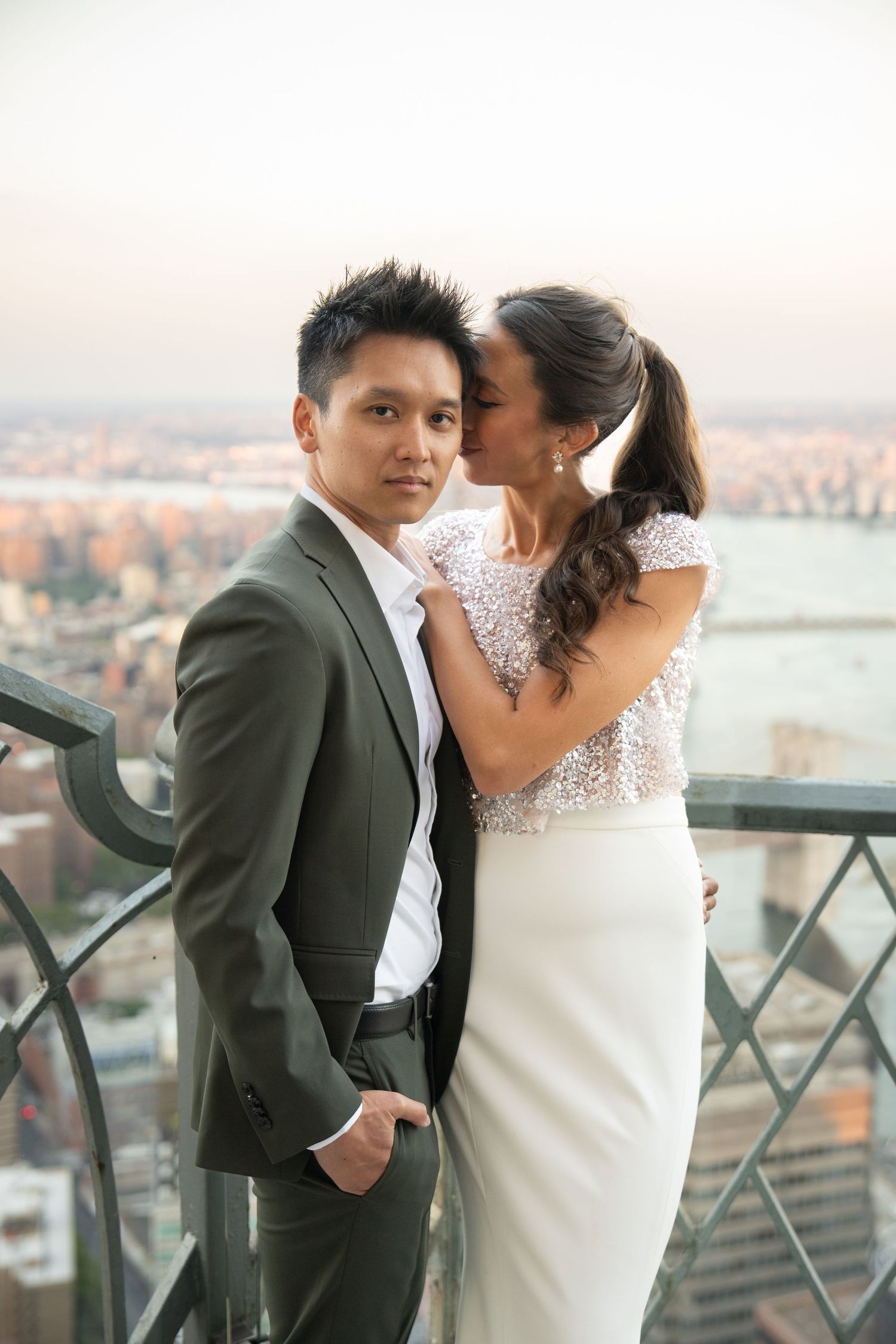 Bride in white sequin dress whispering to groom in dark suit on a city rooftop at dusk.